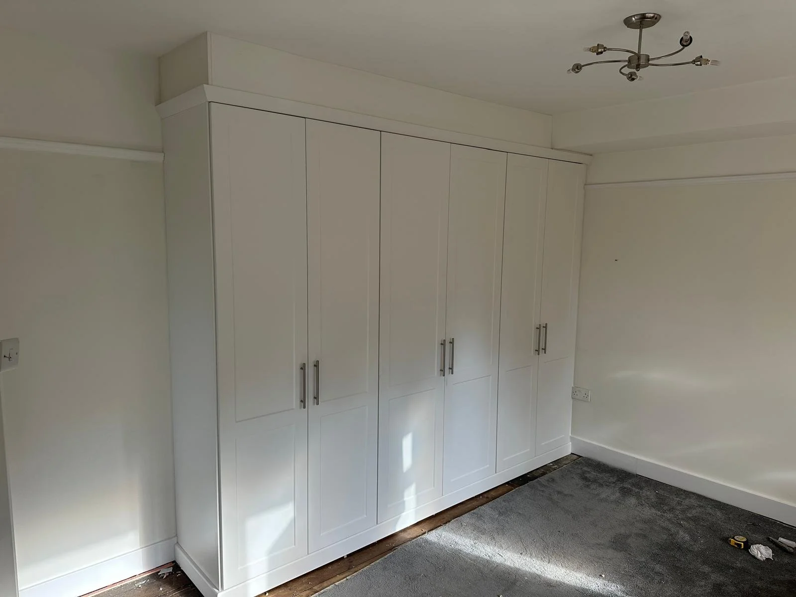 White built-in wardrobe with silver handles in a room with light-colored walls and a gray carpet, with some tools on the floor.