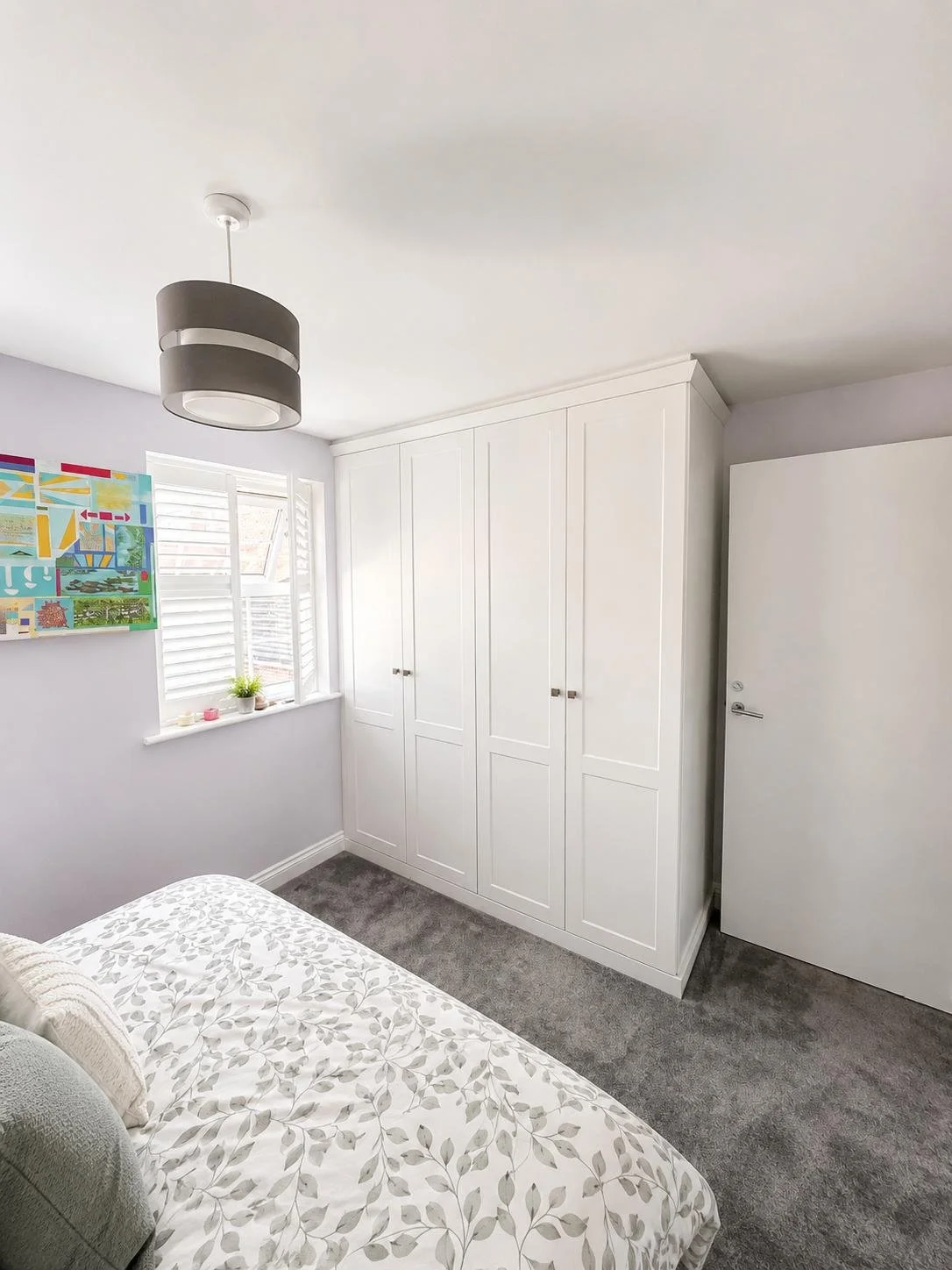 A bedroom with a bed, a window with white shutters, a dresser, floating shelves, and a ceiling light fixture.