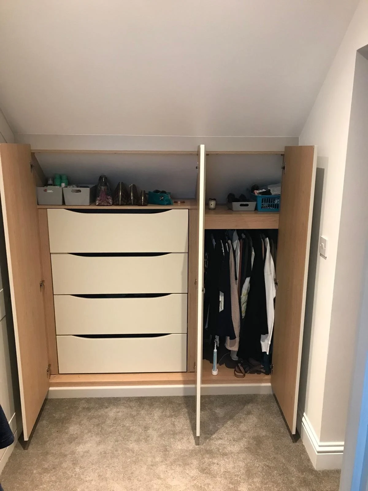 Open wooden wardrobe with four drawers on the left and hanging clothes on the right, set against white walls and carpeted floor.