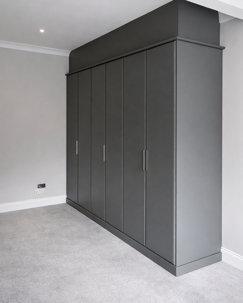 Gray built-in wardrobe with multiple doors in a corner of a room with white walls and light gray carpeted floor.