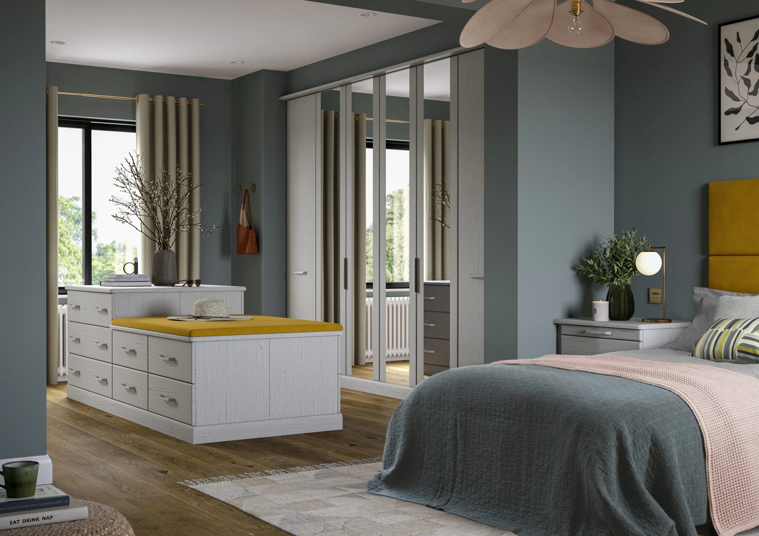 A cozy bedroom with teal walls, a large bed with a gray quilt and patterned pillows, a white dresser with a yellow cushion on it, and a bedside table with a green vase, a lamp, and a book. There's a window with beige curtains, a framed botanical prin