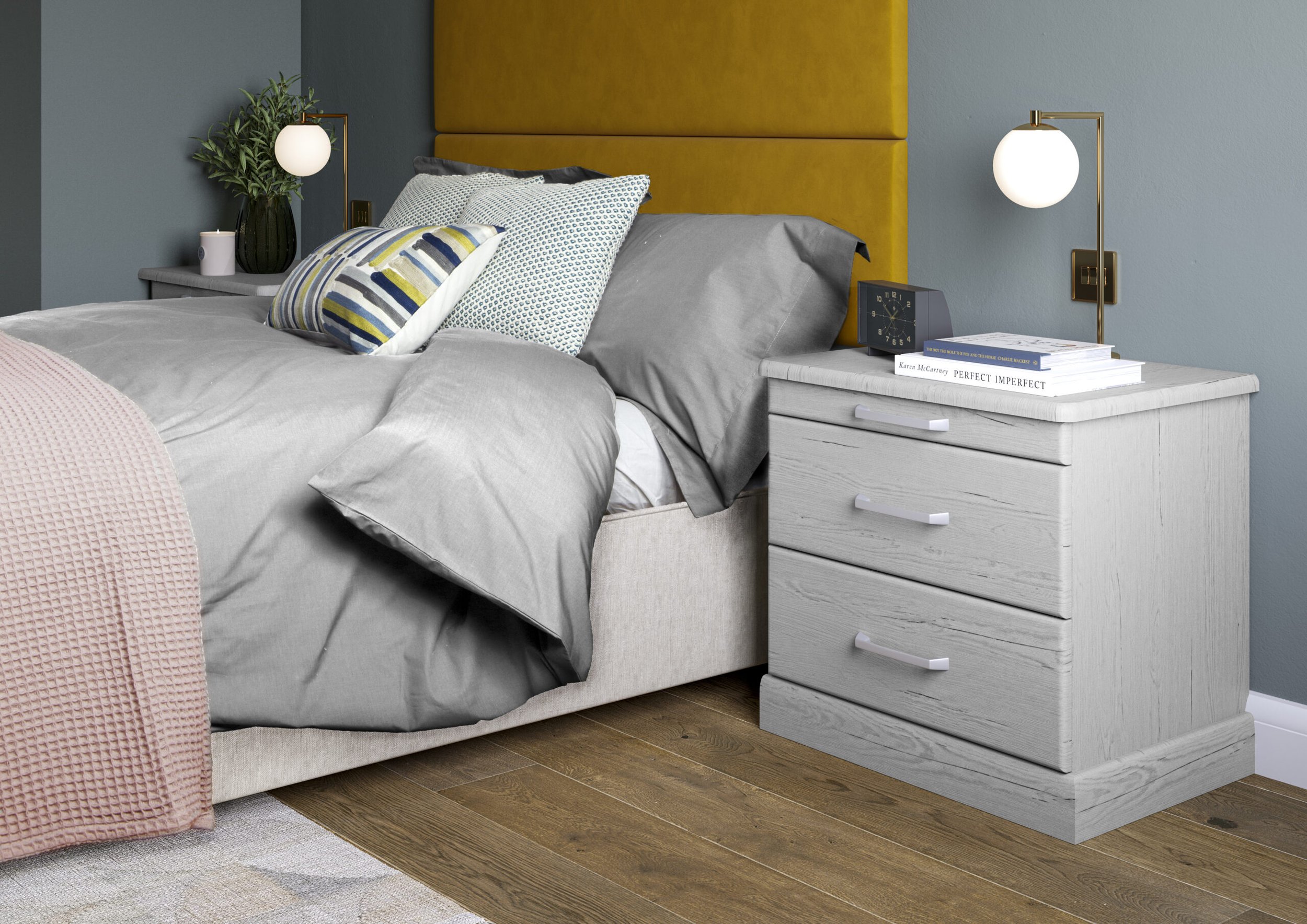 A bedroom with a beige upholstered bed with a tan headboard, gray bedding, and assorted decorative pillows. A white nightstand with a stack of books and a blue clock, a black vase with green plants, and a pink blanket over the side of the bed. Two wa