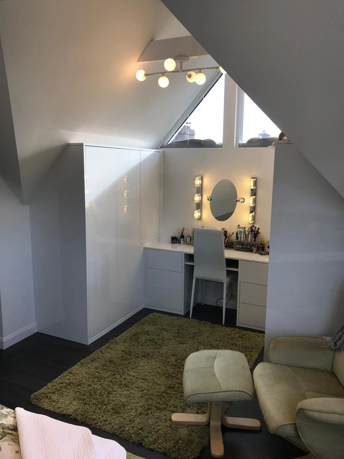 A cozy attic room with a white vanity desk, mirror, and lighting, a beige armchair and ottoman, a green shaggy rug, and a sloped ceiling with a skylight window.