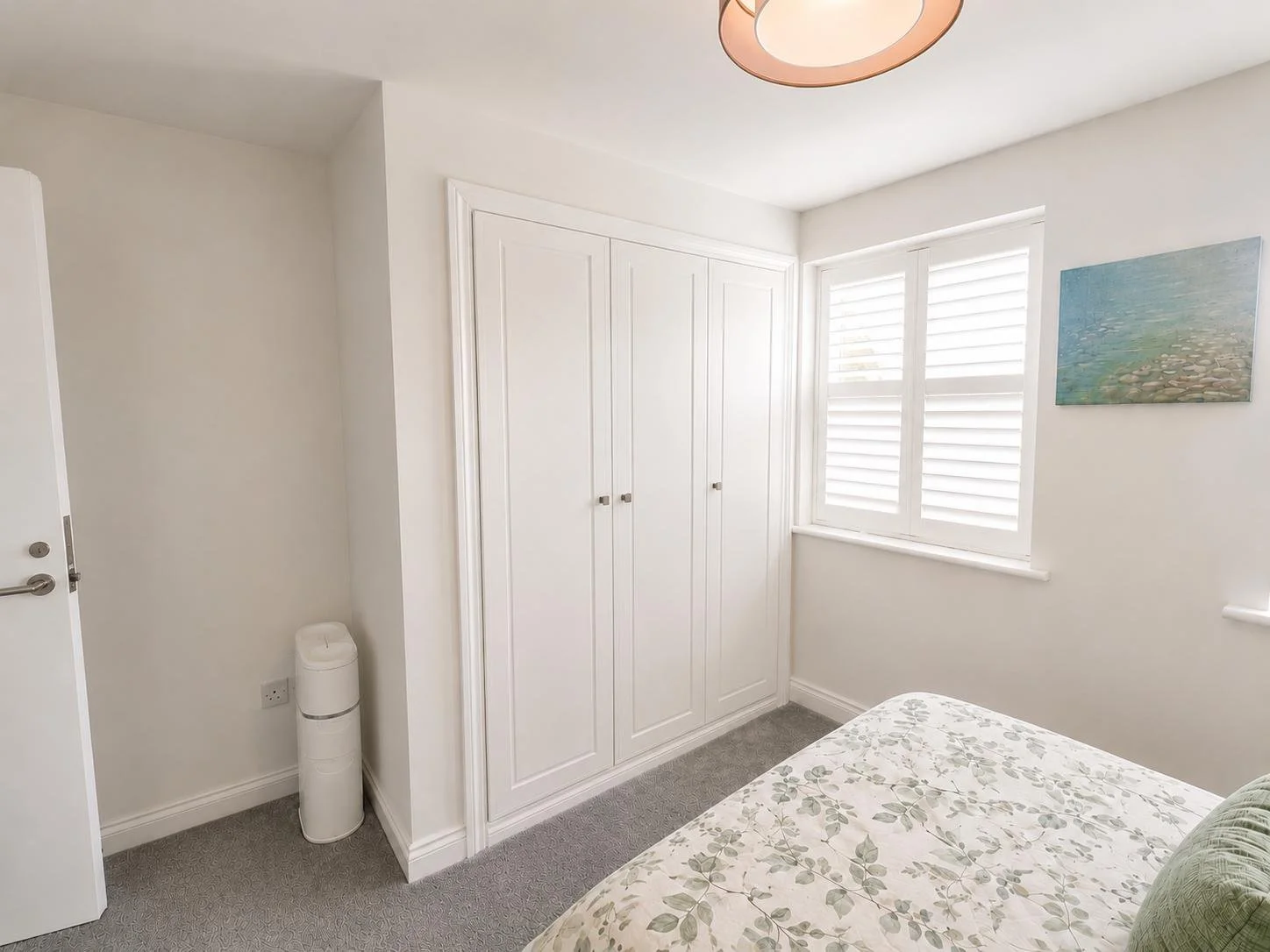 A bedroom corner with a white built-in wardrobe, window with white shutters, a bed with floral-patterned bedding, a painting on the wall, and a small white trash can in the corner.
