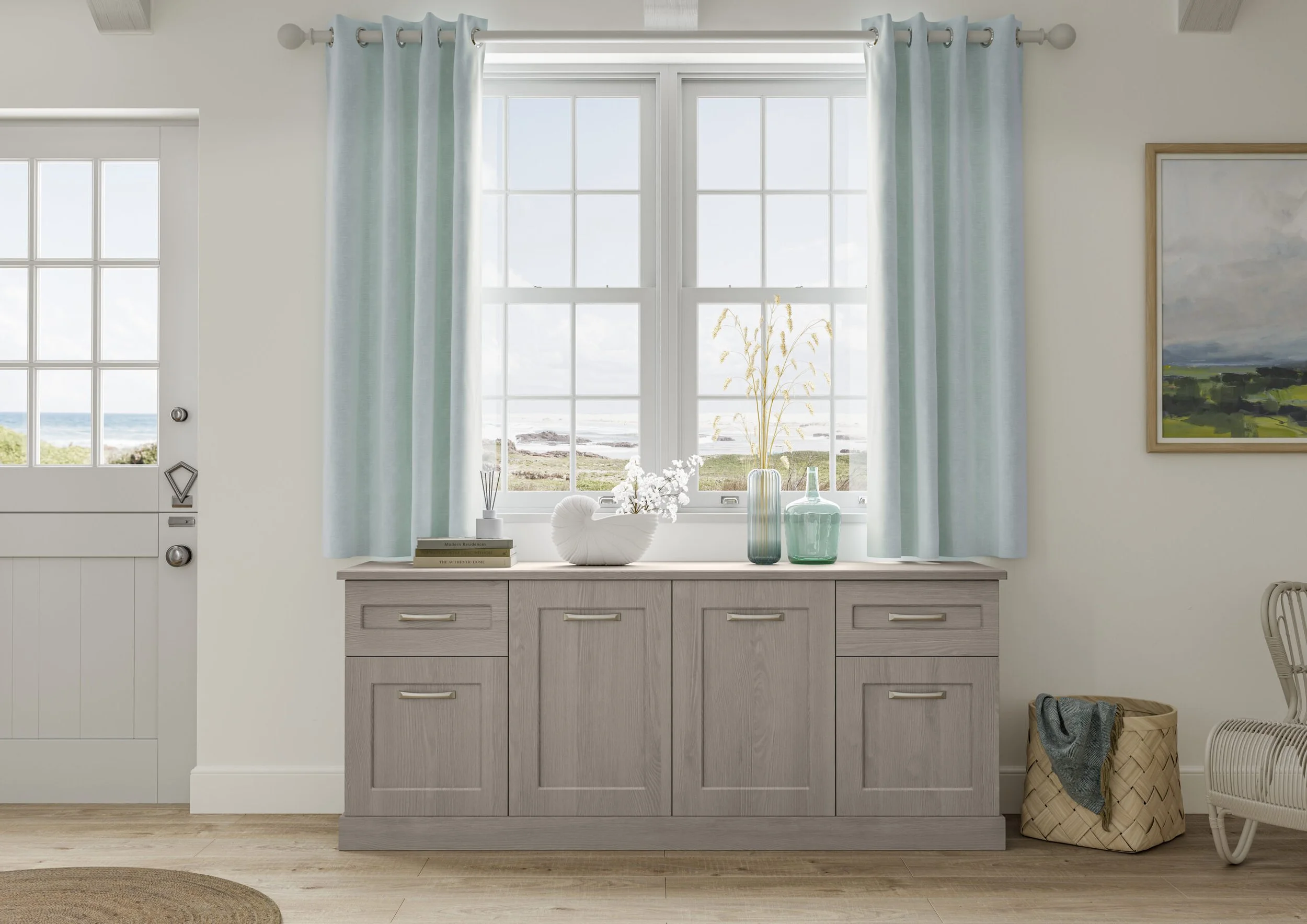 A kitchen interior with a large window overlooking a beach, light blue curtains, a wooden sideboard with decorative vases and flowers, and a basket with a towel.