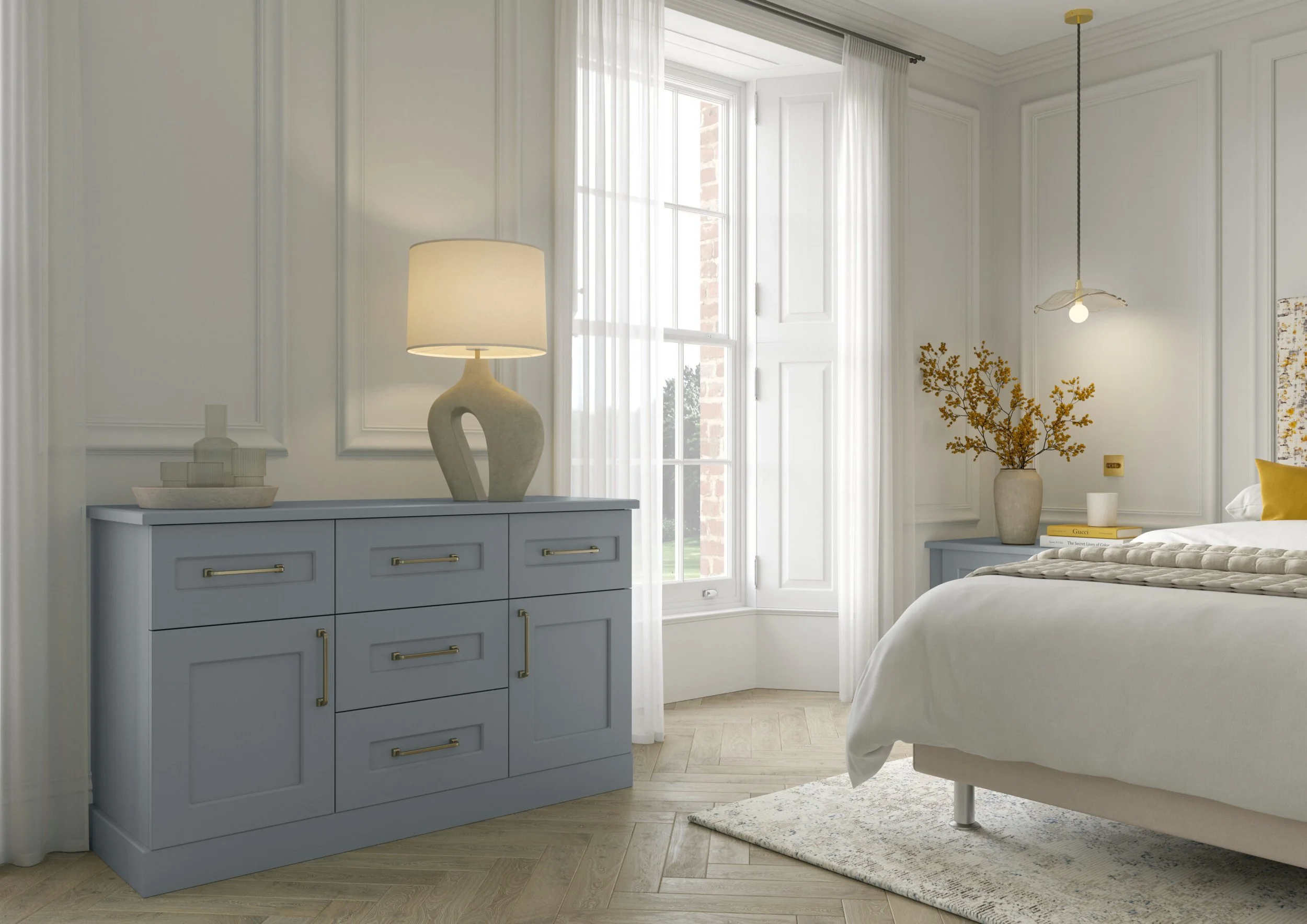 A bedroom with a light blue dresser, a lamp with a sculptural base, a large window with white curtains, a bed with white bedding, a vase with yellow flowers, a stack of books, and a modern pendant light fixture.