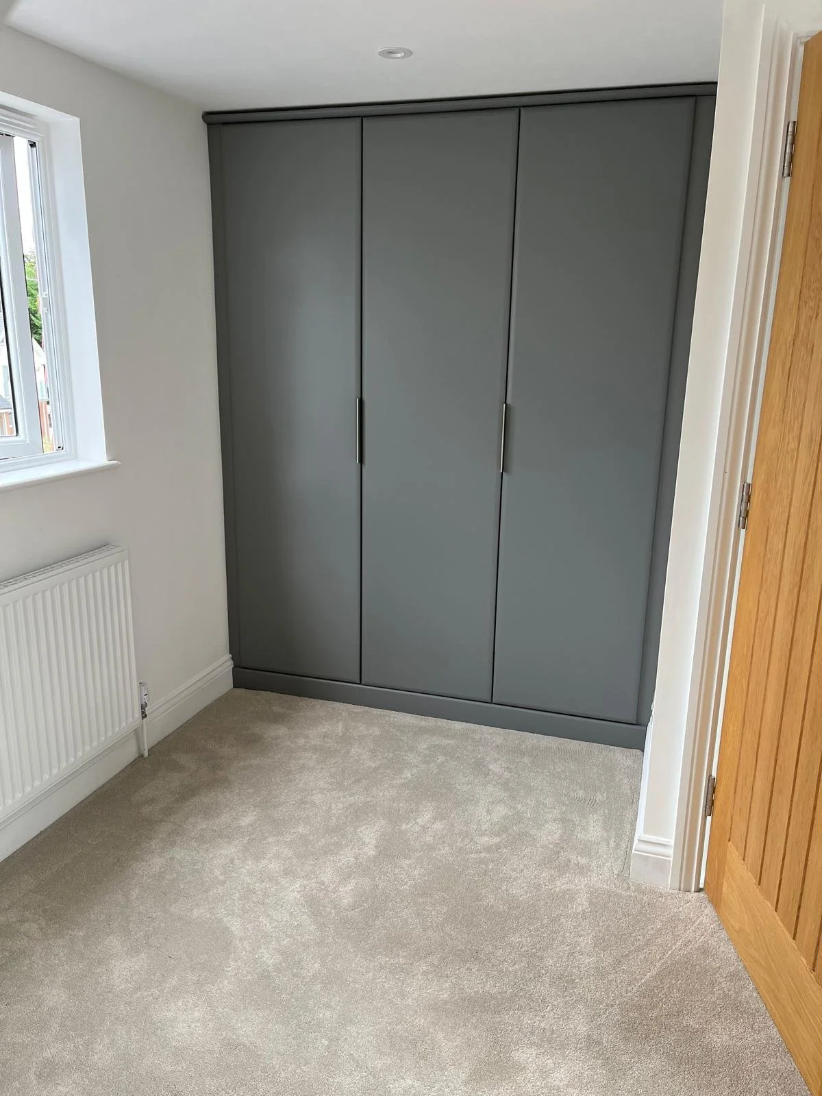 Gray wardrobe with three doors in a corner of a room with beige carpet, a window, white walls, and a wooden door.
