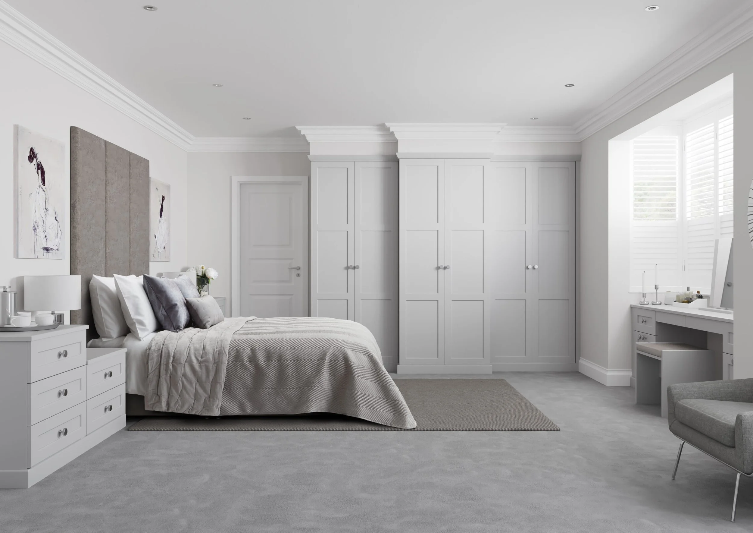 A bedroom with a white bed, gray bedding, and pillows, flanked by white nightstands with lamps. On the wall, there are two art pieces and a large gray headboard. The room has white cabinetry, crown molding, a window with shutters, and a gray carpet.