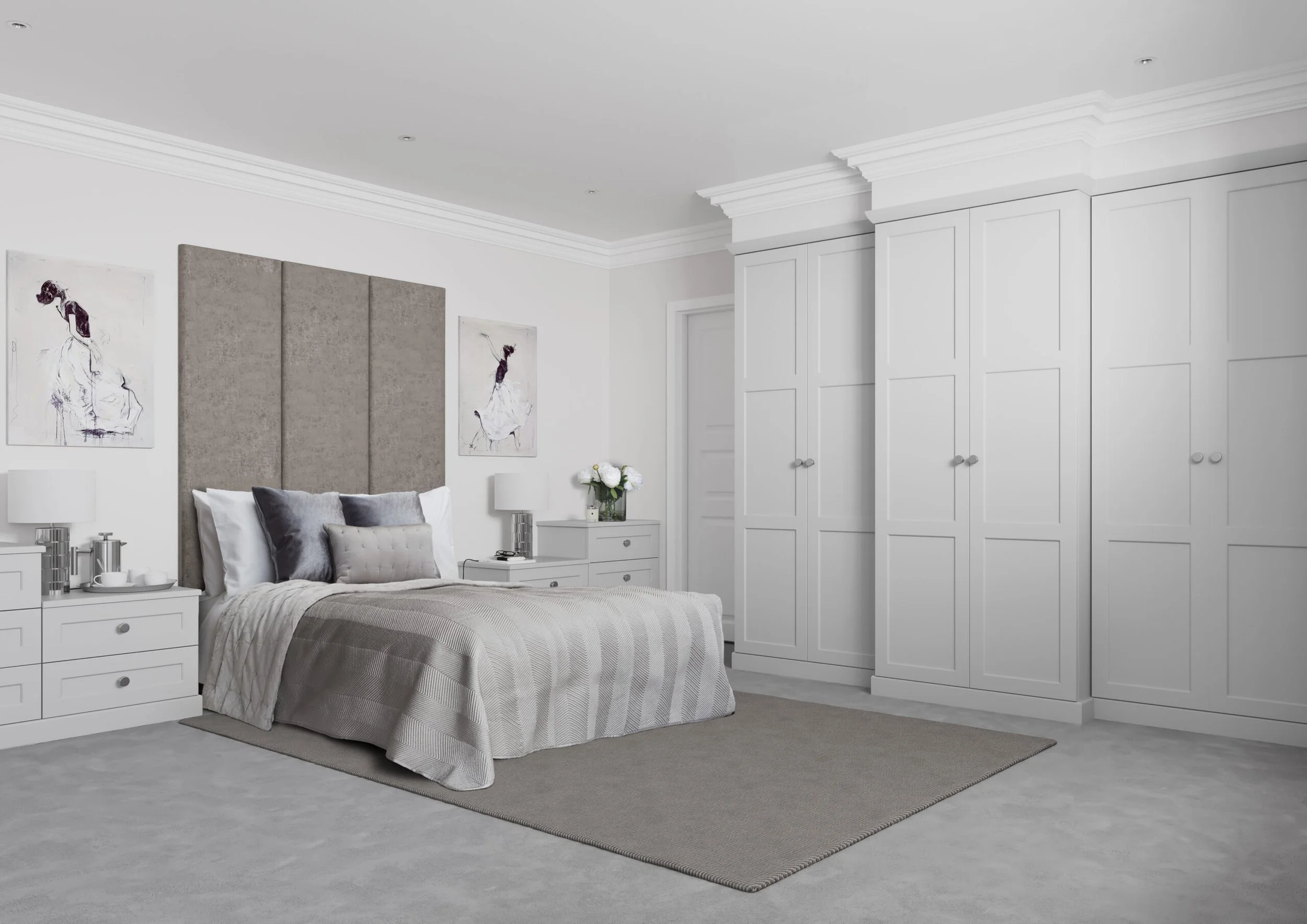 A modern bedroom with a white color scheme, featuring a bed with a beige tufted headboard, white bedding, and throws in shades of gray. On either side of the bed are white nightstands with lamps. Two framed black-and-white fashion illustrations hang on the wall above the bed. There are white built-in wardrobes and a small table or dresser with a vase of white flowers. A light gray area rug covers part of the plush carpeted floor.