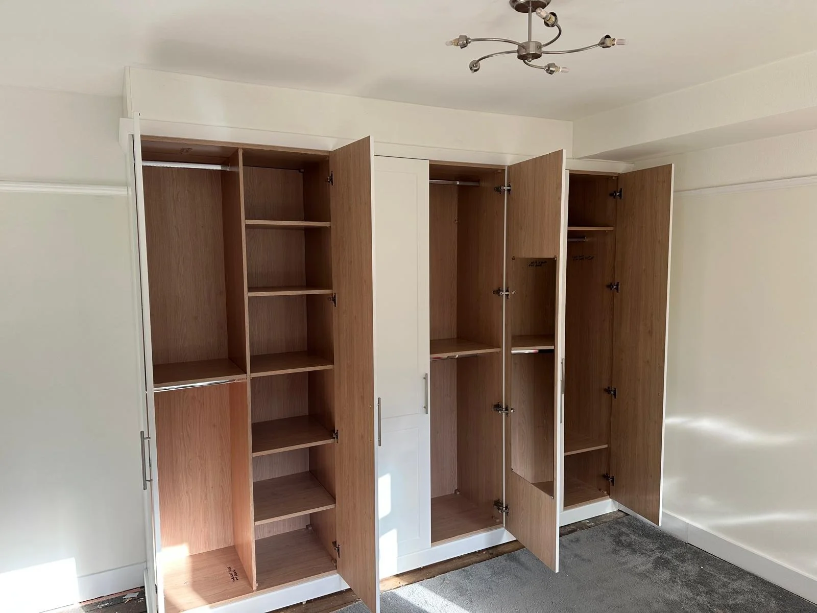 Open wooden wardrobes with multiple shelves and hanging rods in a room with off-white walls and a modern ceiling light fixture.