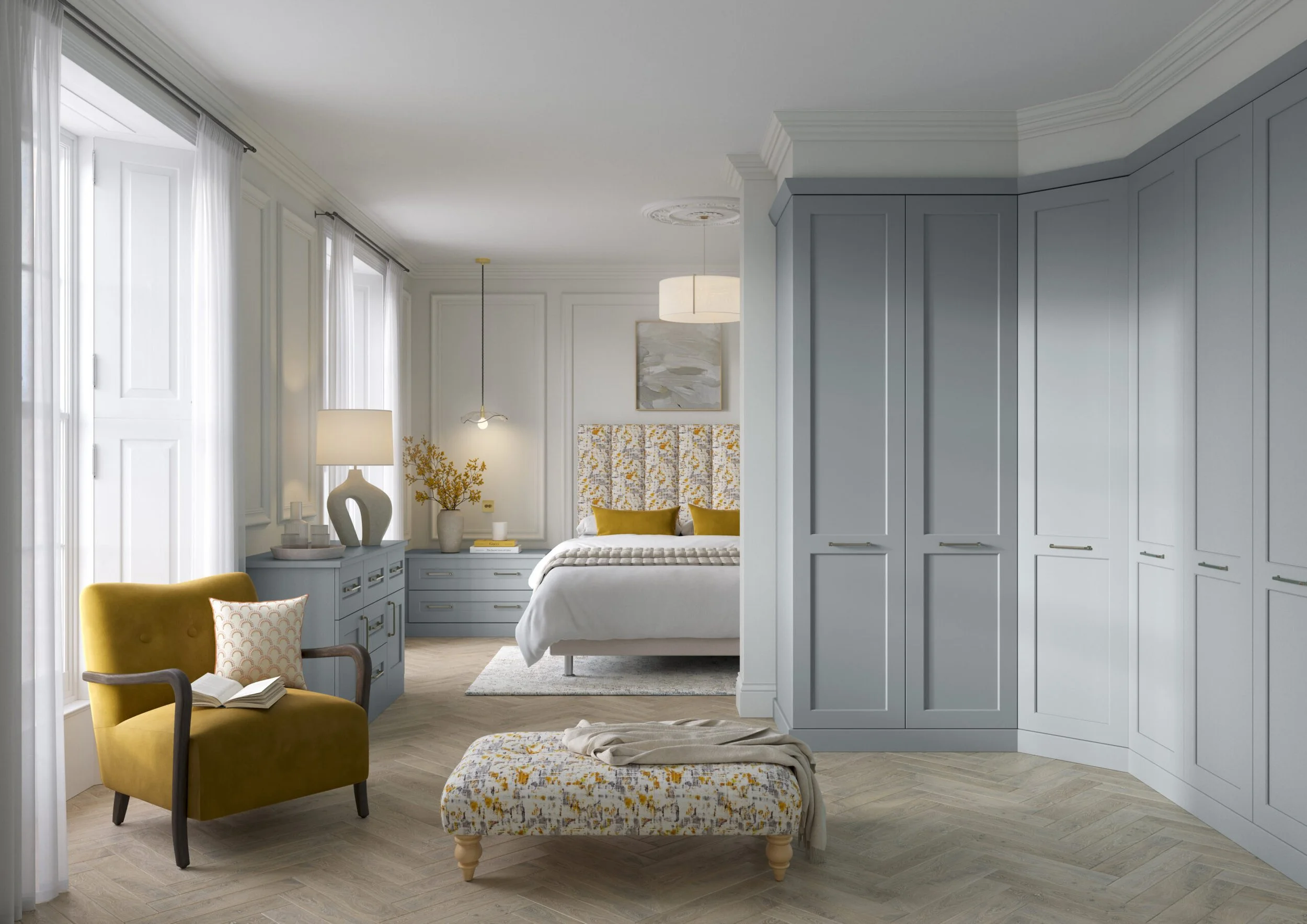 A bright bedroom featuring white curtains, a yellow armchair with a patterned pillow, a nightstand with a white vase and flowers, a bed with white bedding and yellow pillows, abstract artwork on the wall, and a large built-in gray wardrobe.
