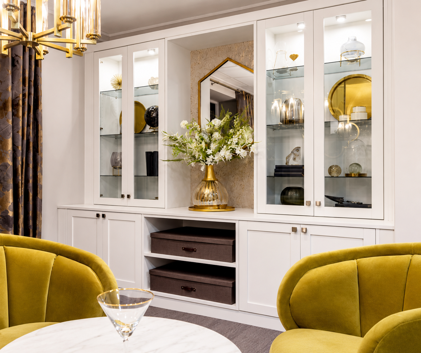 A decorated living room with a white built-in cabinet displaying decorative objects, a large floral arrangement in a gold vase, and yellow chairs around a white marble table, with a gold chandelier and patterned curtains.