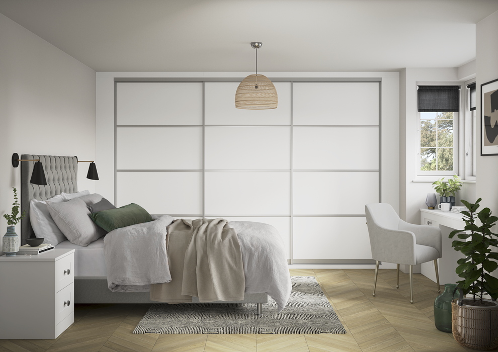 A modern bedroom with a white bed, gray headboard, green throw pillow, cream blanket, and a white nightstand. There is a bed with a bedside lamp, a white armchair, a large green houseplant, and a window with a view of trees.