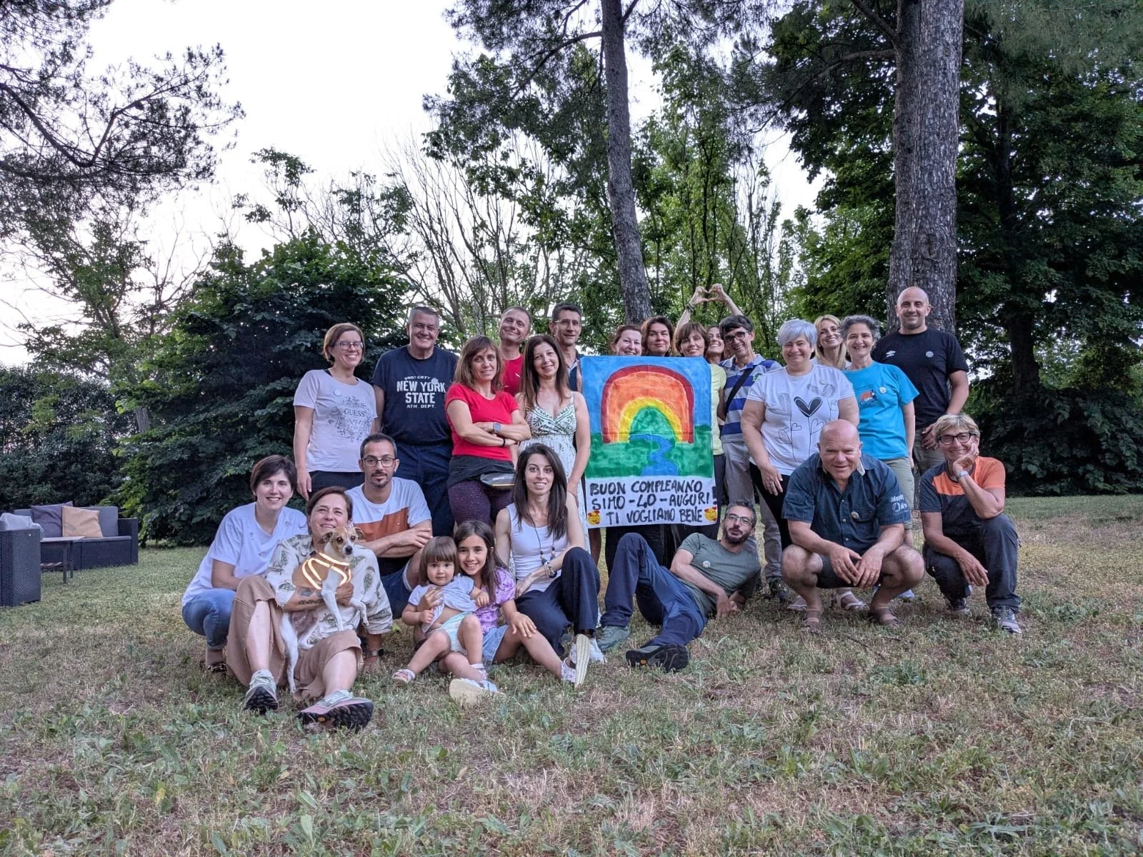 Foto di gruppo all'aria aperta con alberi sullo sfondo. I partecipanti sorridono e mostrano un cartellone disegnato con un grande arcobaleno.