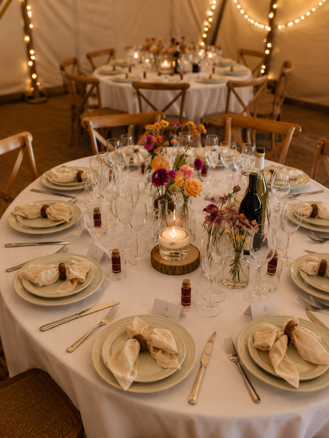Elegant banquet table with white tablecloth, floral centerpieces, lit candle, wine bottles, and place settings with napkins, glasses, and silverware. String lights adorn the background.
