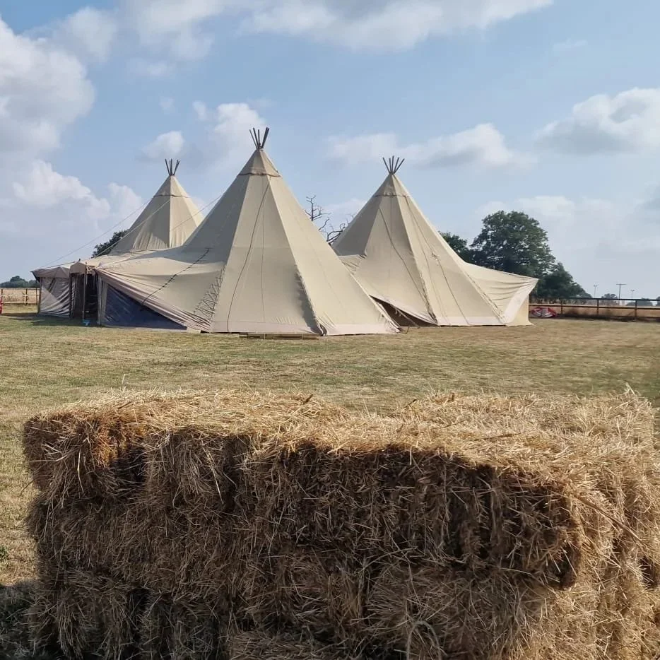 Three teepee-style tents set up on a grassy field with a hay bale in the foreground and trees and a partly cloudy sky in the background.