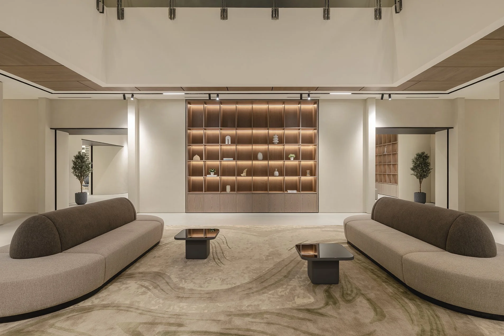 A modern, minimalistic lobby or lounge with two curved beige sofas facing each other, small black coffee tables, two potted plants, and a large wooden shelving unit with decorative items and books behind. The ceiling features wooden panels and modern lighting.