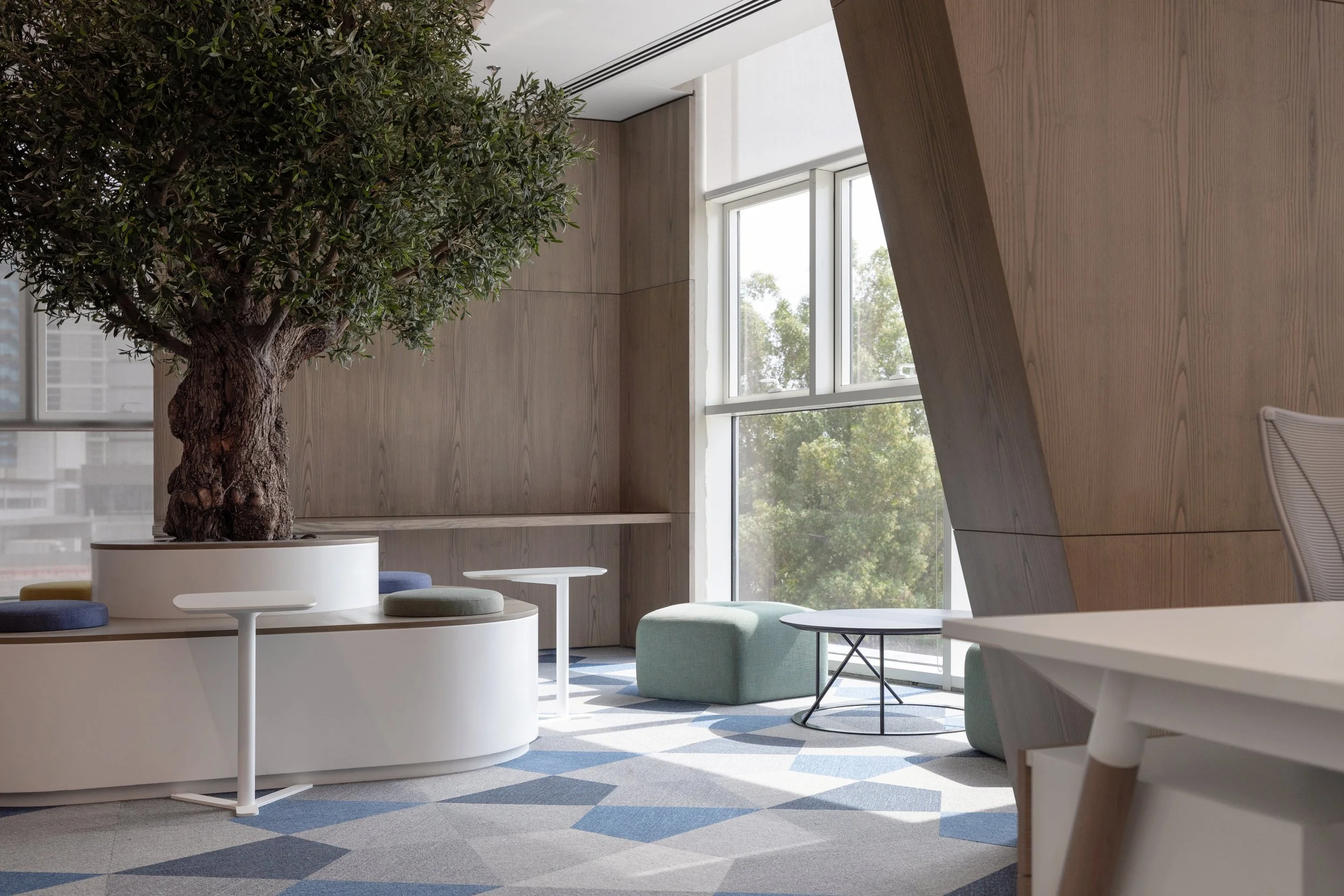 Modern interior space with wooden walls and large windows. Features a potted tree, white and pastel-colored seating, round tables, and a patterned carpet.