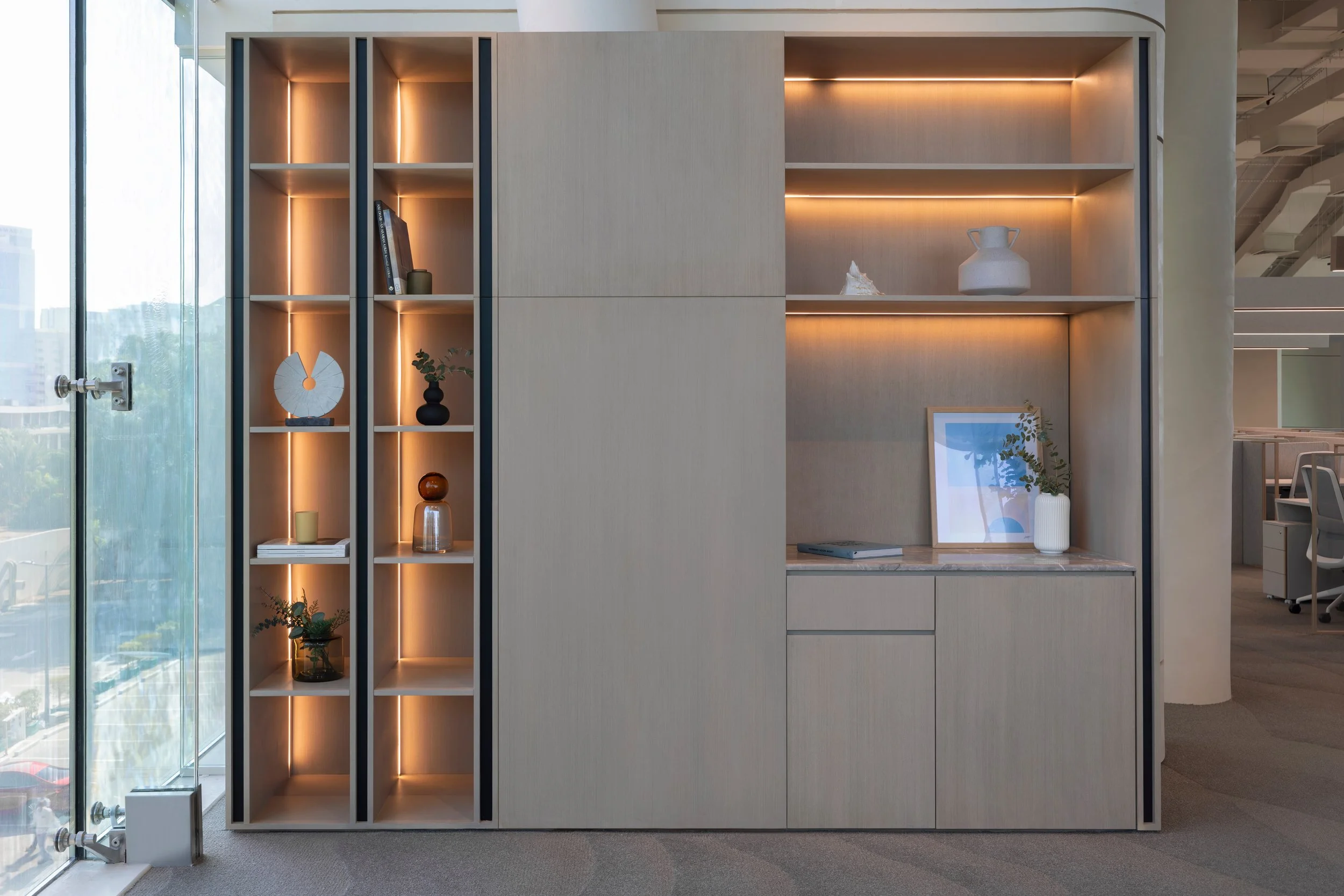 Modern beige bookshelf with open and closed compartments in an office setting, decorated with vases, books, and artwork, with large windows and cityscape view in the background.