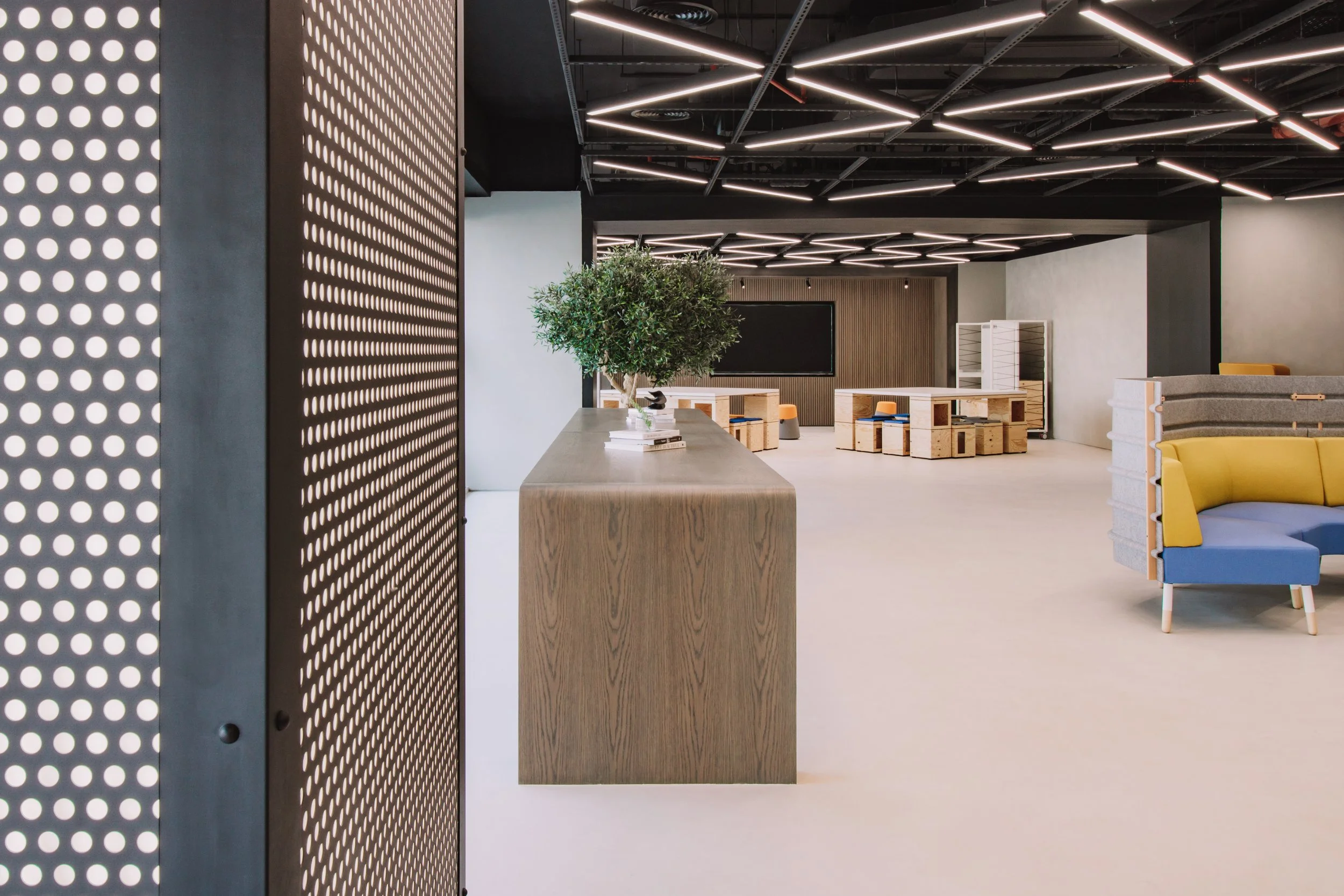 Modern office interior with a perforated metal divider, a long wooden table, a potted plant, and contemporary furniture with colorful cushions, illuminated by geometric LED ceiling lights.