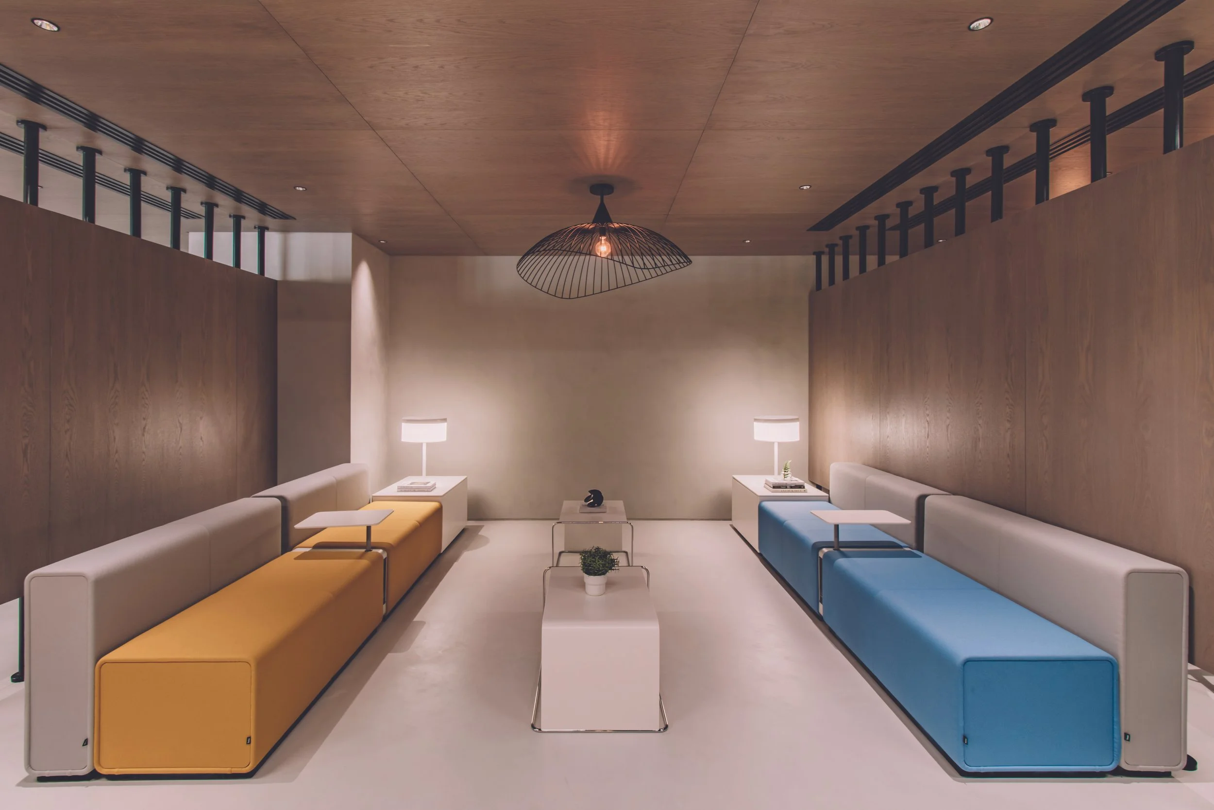 Modern minimal living room with beige and blue sofas, white side tables, a coffee table with a potted plant, wood-paneled walls, a wooden ceiling, a centrally hanging black wire pendant light, and two white table lamps.