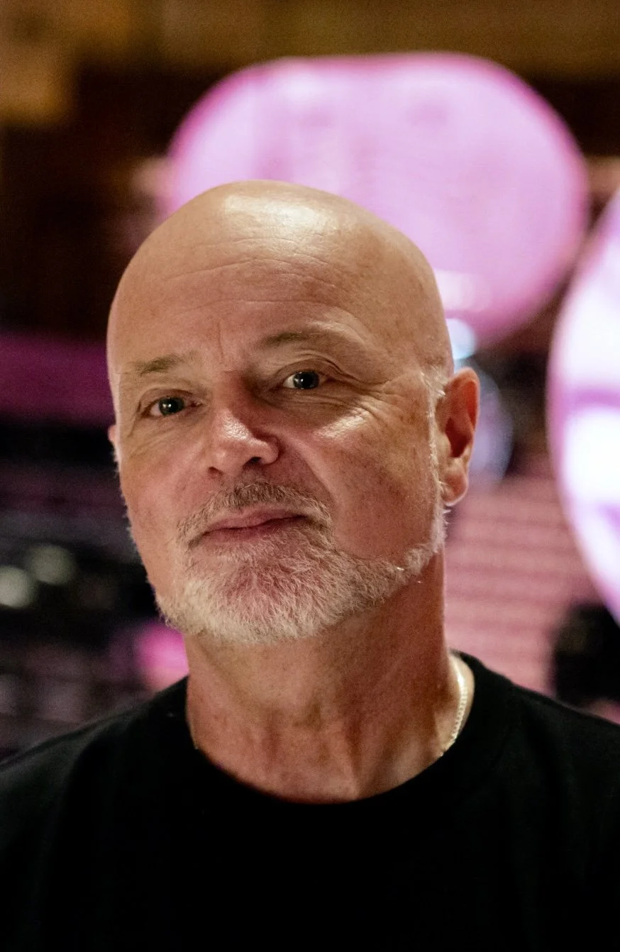 A close-up of a bald man with a beard, wearing a black shirt, in front of blurred pink lights.
