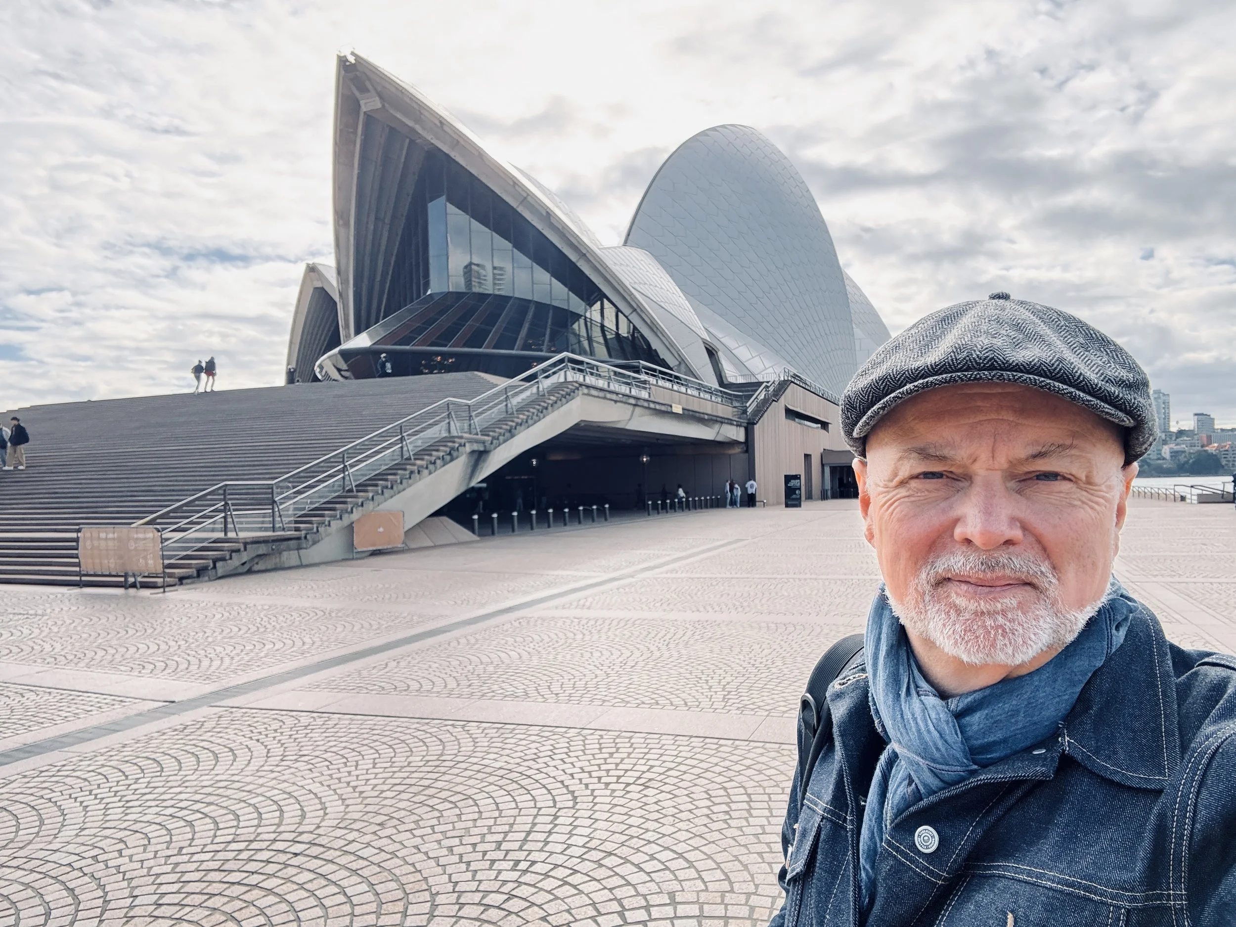 A man with a cap and denim jacket taking a selfie in front of the Sydney Opera House with a cloudy sky background.
