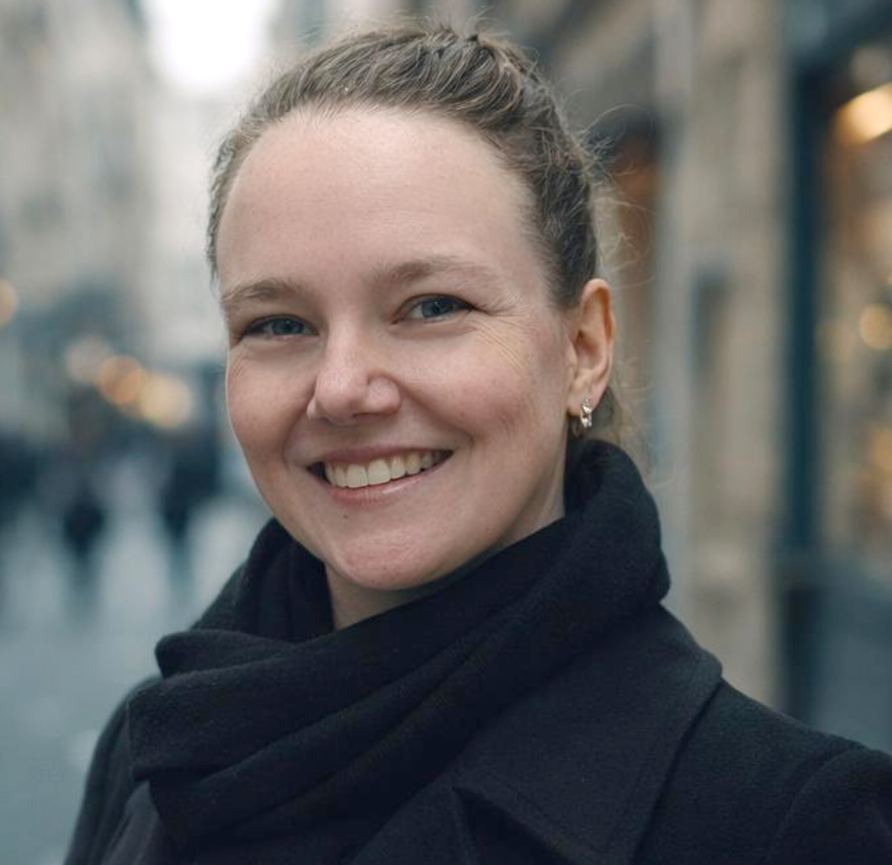 Close-up of a woman with a smiling expression, wearing earrings and a black scarf, with blurred lights and urban background.