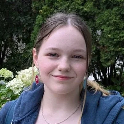 Young woman with long hair and earrings, outdoors with green trees and white flowers in the background.