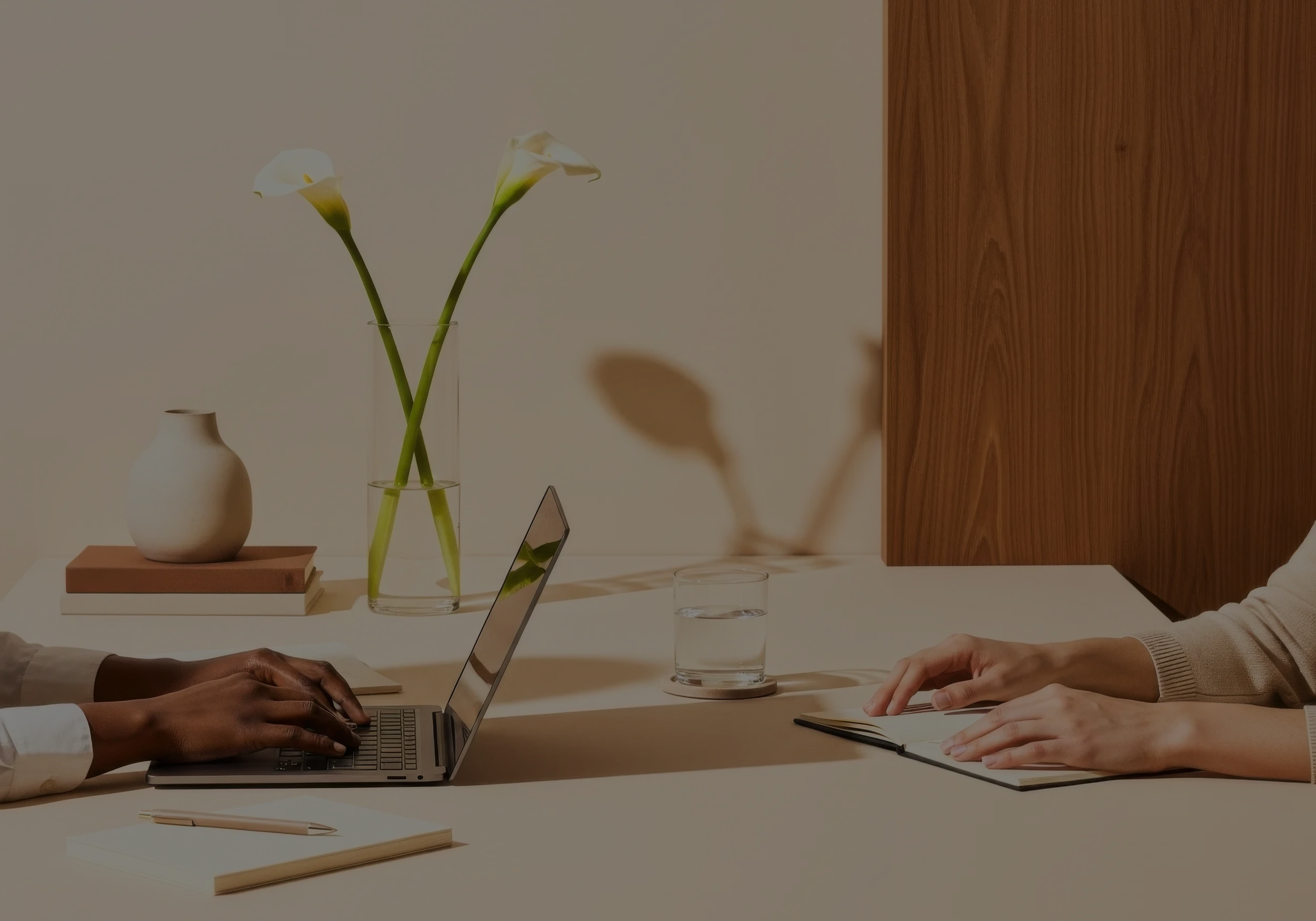 Two people sitting at a table with laptops, notebooks, and pens, having a discussion. There are decorative vases with white calla lilies and glasses of water on the table, against a beige wall with a wooden panel.