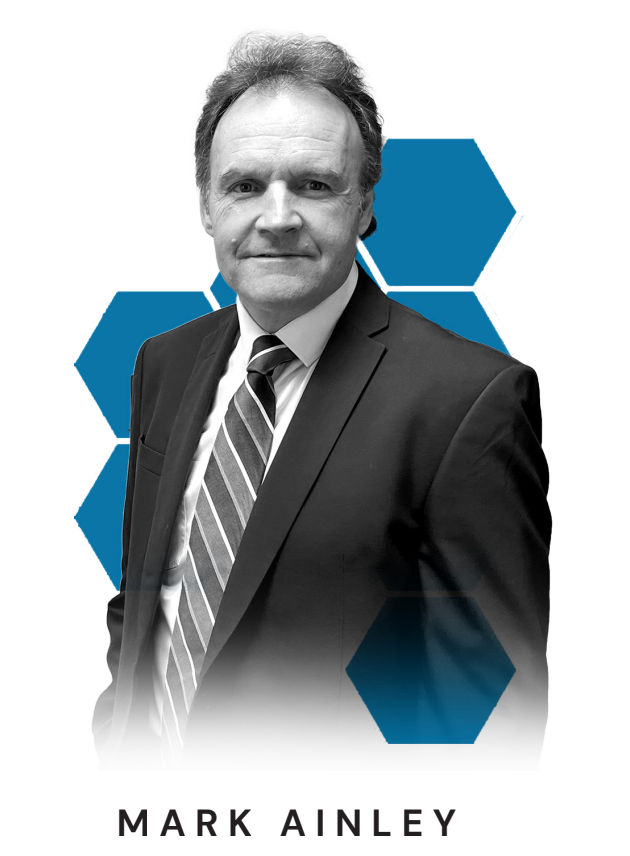 Black and white portrait of Mark Ainley in a suit with a striped tie, standing in front of a background with blue hexagons.