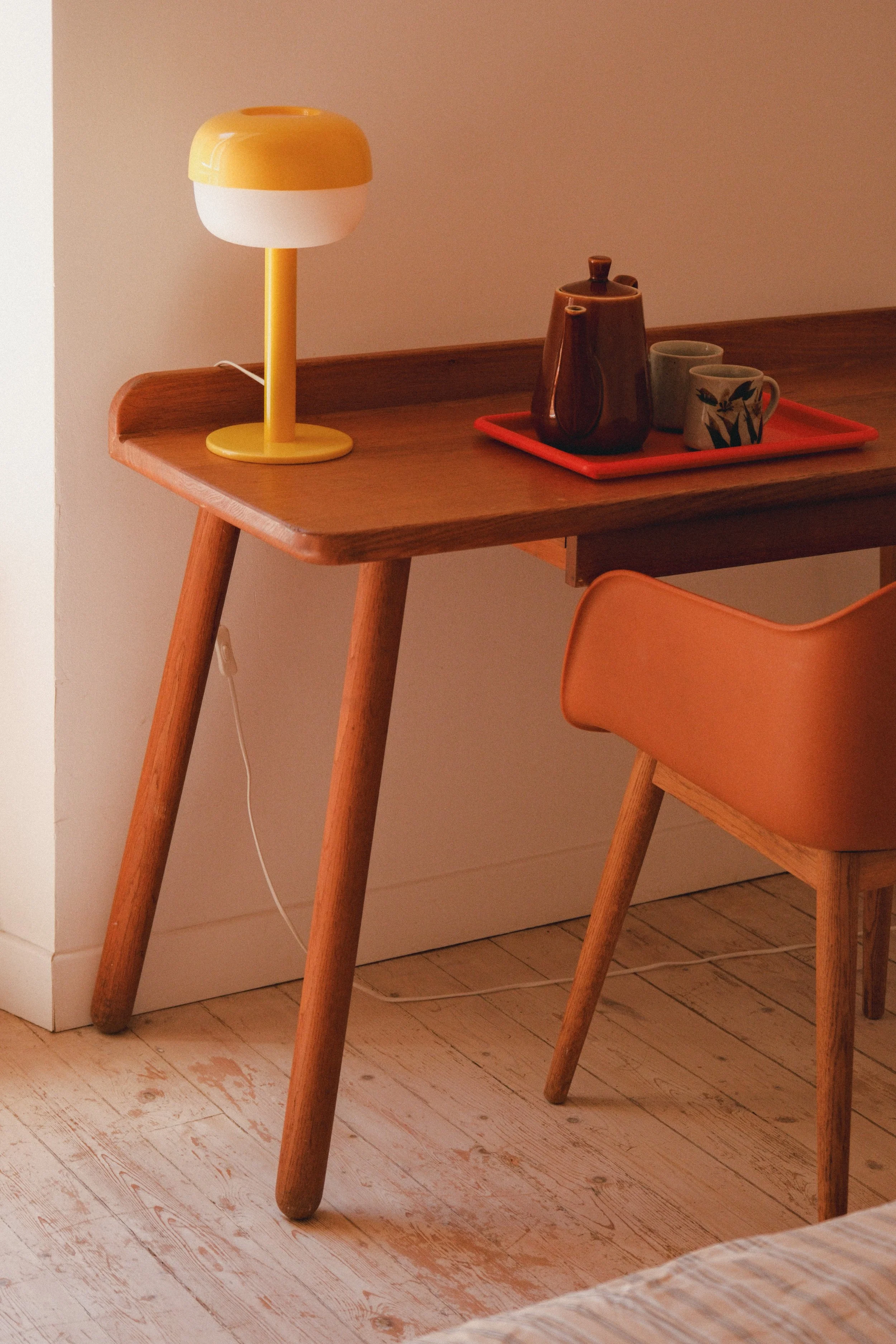 Une table en bois avec une lampe jaune et blanche, une carafe marron, deux tasses et un plateau rouge.