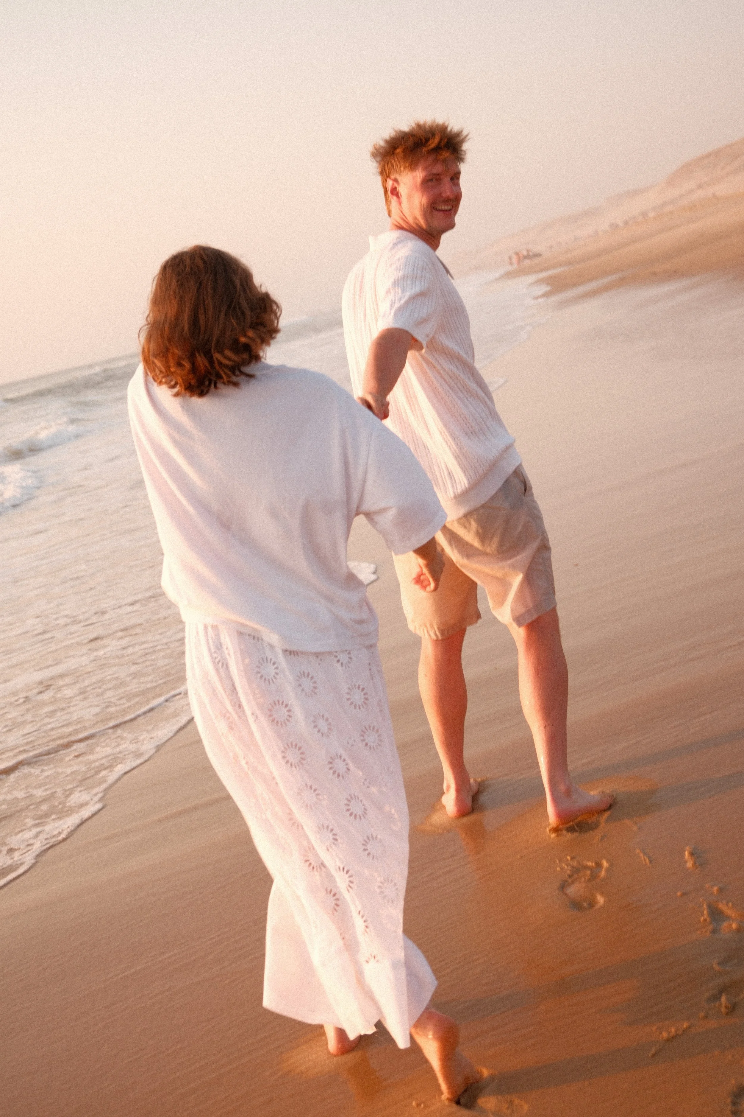 Deux personnes, un homme et une femme, marchent pieds nus sur la plage au coucher du soleil. L'homme regarde en arrière avec un sourire, tandis que la femme marche devant eux. Le sable, la mer et une falaise en arrière-plan complètent la scène tranqu