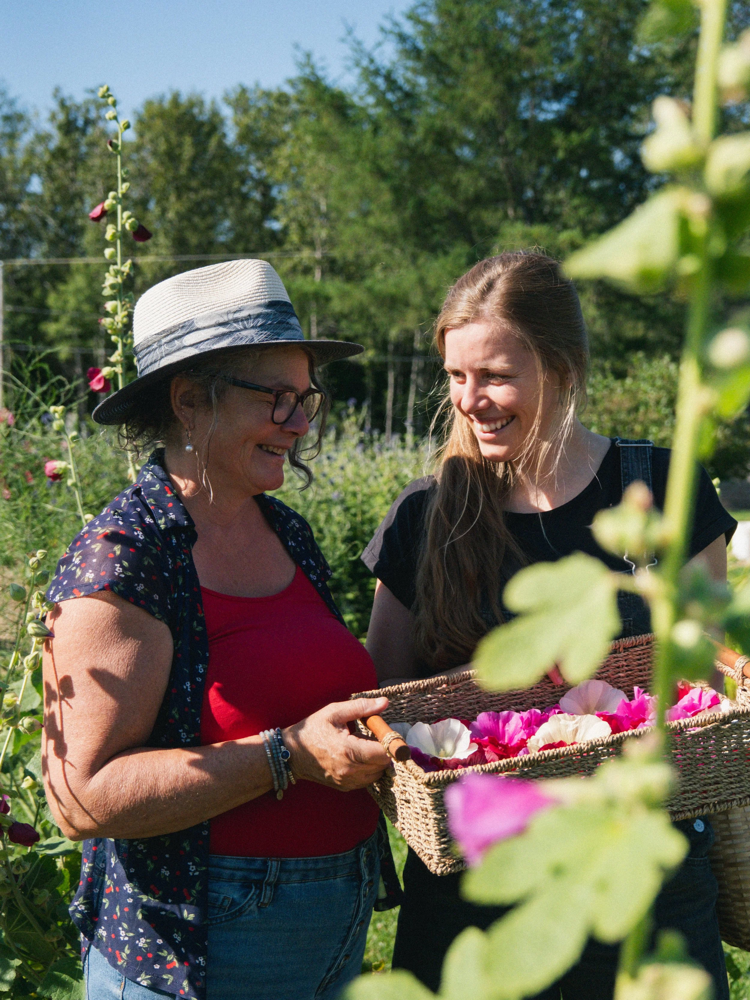 Deux femmes souriantes dans un champ de fleurs, l'une tenant un panier rempli de fleurs colorées.