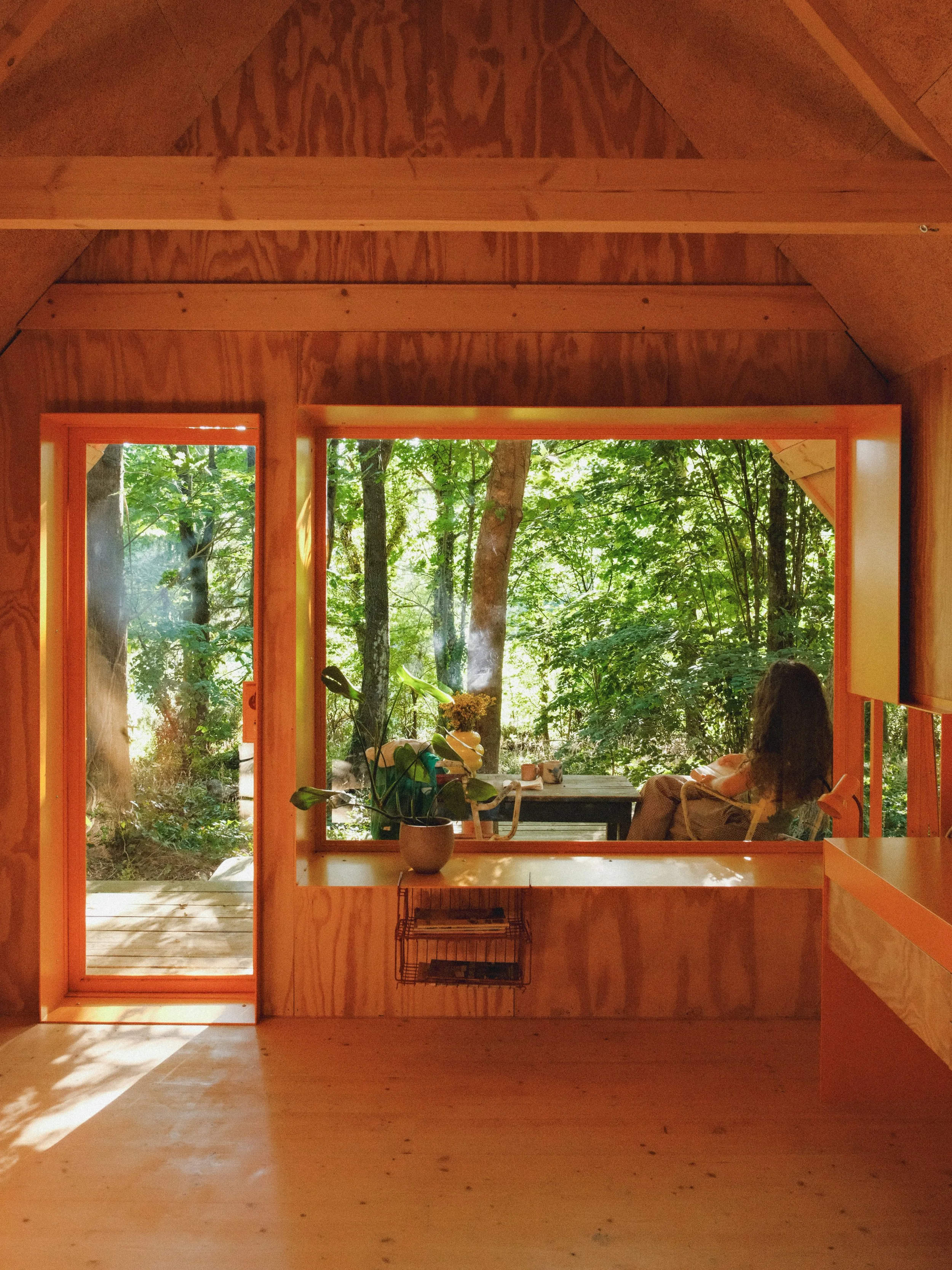 Une pièce en bois avec une grande fenêtre ouverte sur la forêt, une personne assise sur une chaise devant la fenêtre, il y a une plante en pot sur une table.
