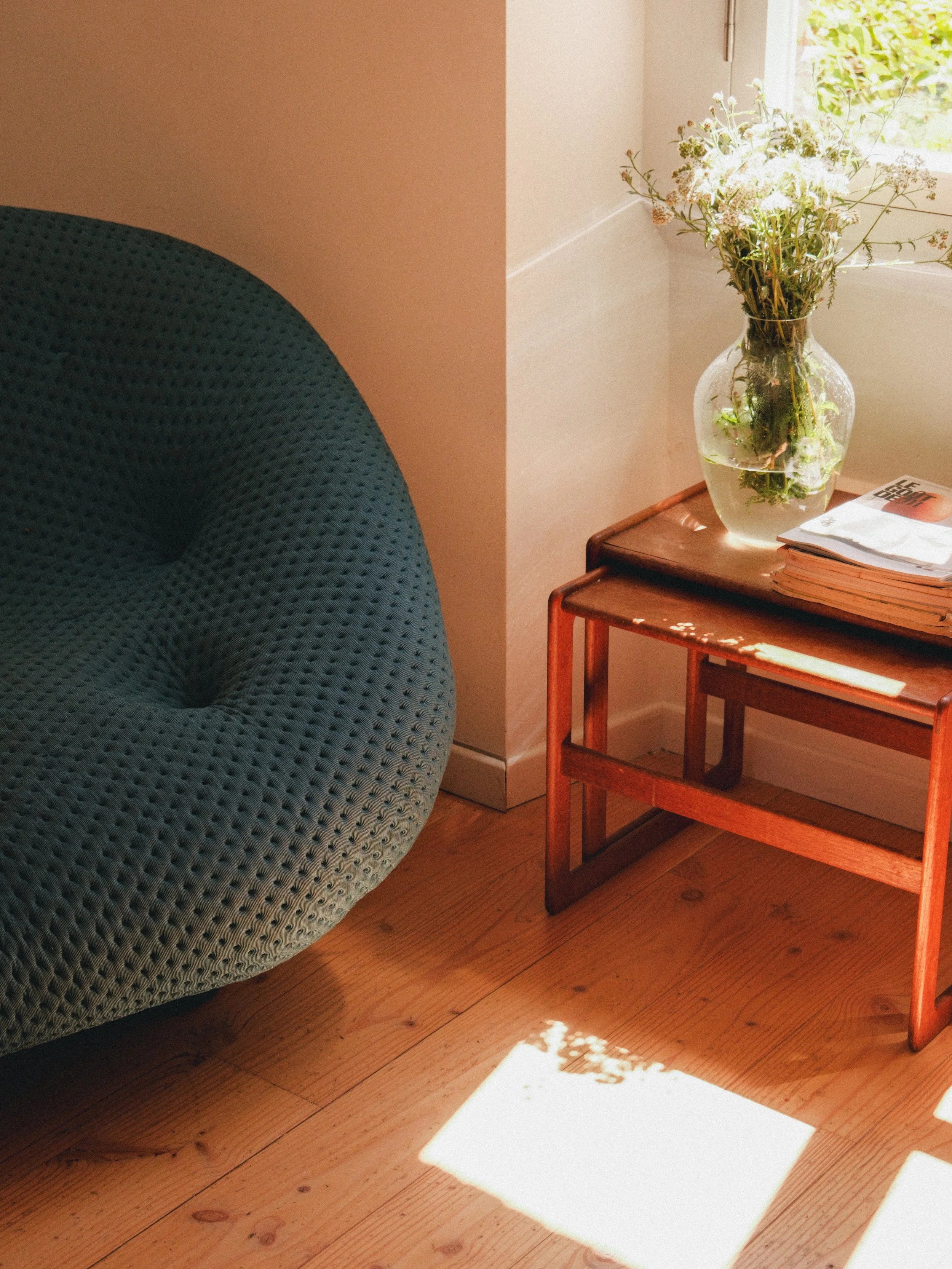 Une plante en vase clair sur une petite table en bois, à côté d’un fauteuil bleu en tissu sur un sol en bois, avec de la lumière naturelle entrant par une fenêtre.