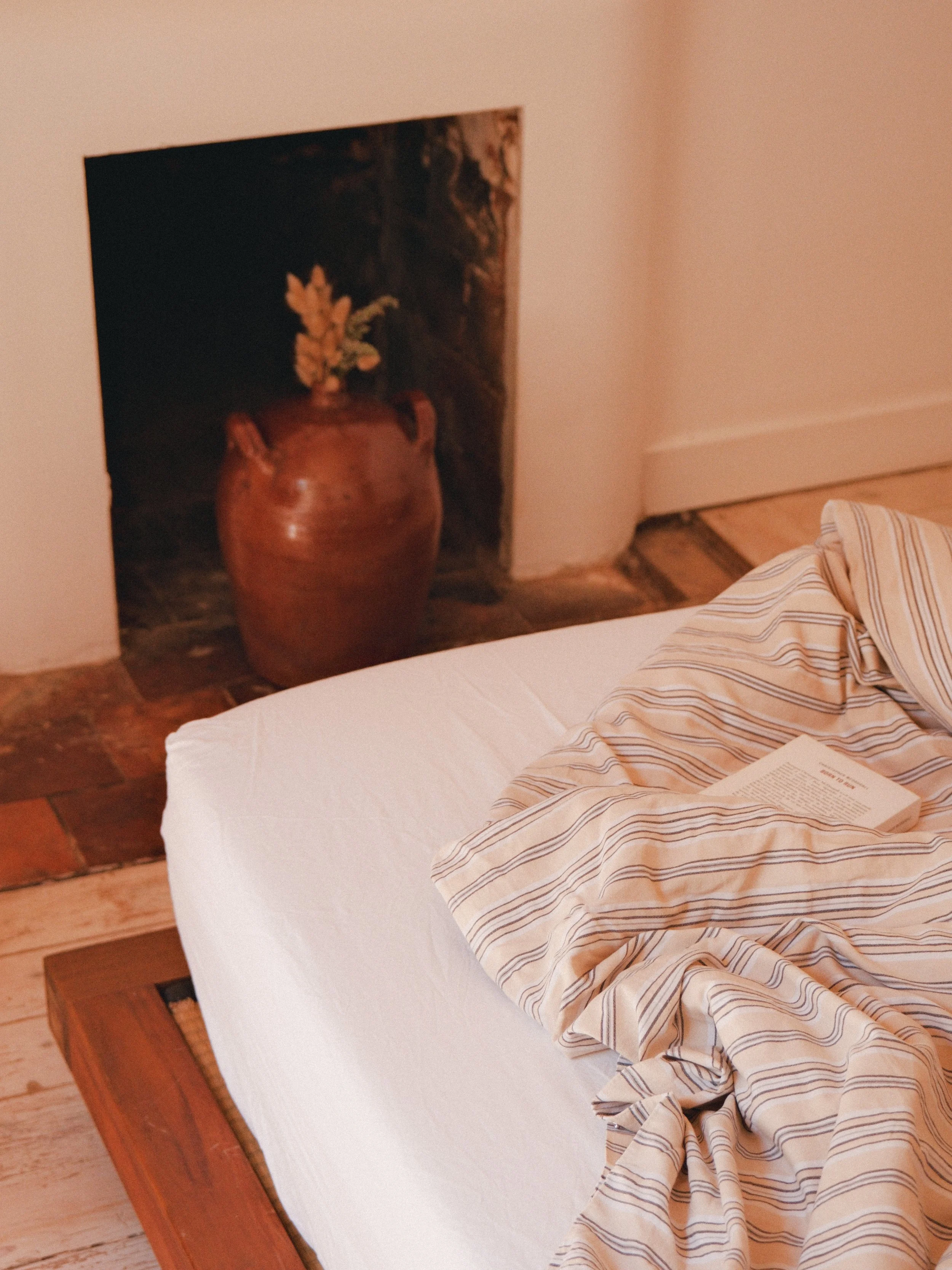 Un lit avec des draps à rayures beige, une serviette ou une chemise à rayures, et un livre posé dessus. En arrière-plan, une grande jarre en terre cuite contenant une plante, dans une pièce avec un mur beige et un sol en bois.