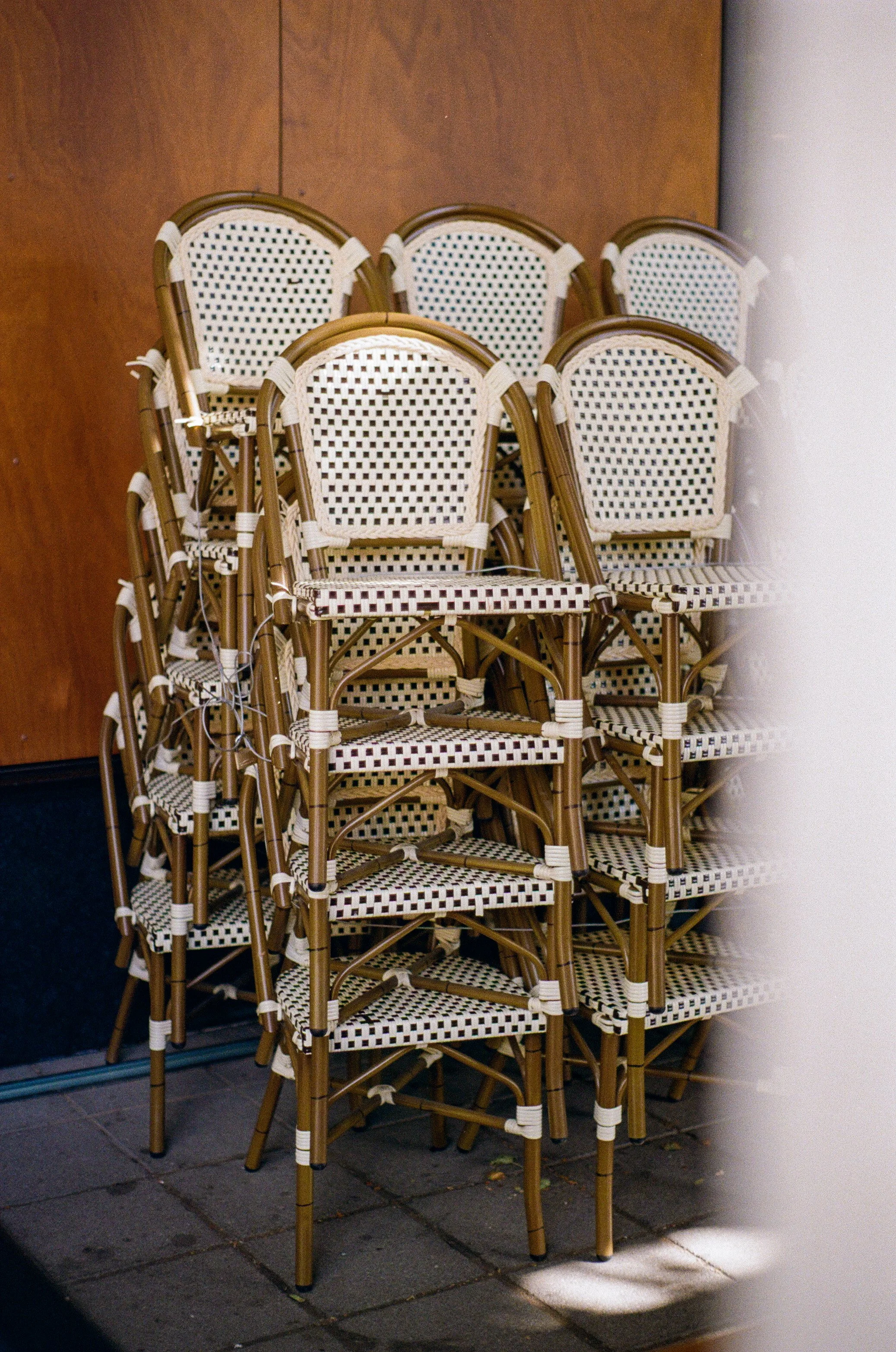 Une pile de chaises en rotin avec assises et dossiers en osier blanc, empilées contre un mur en bois, au pied d'une surface extérieure pavée.