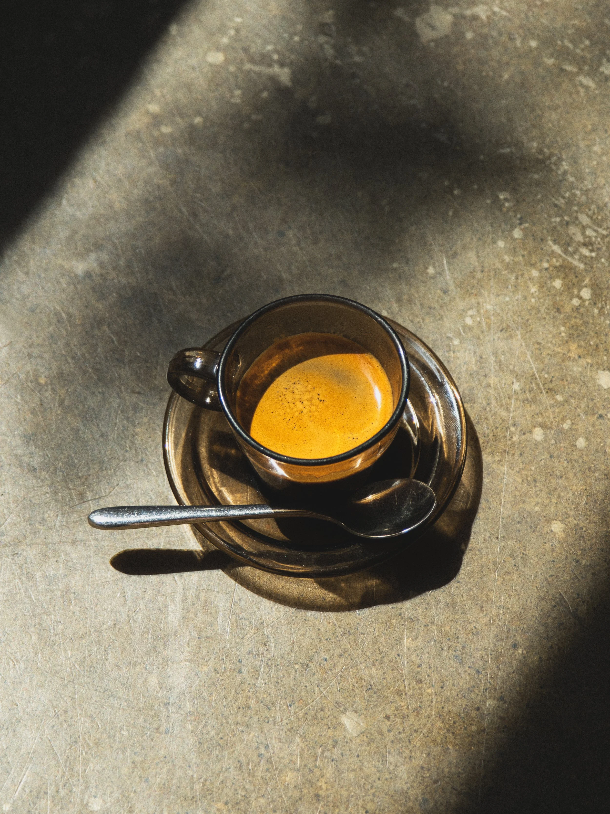 Une tasse de café espresso vue du dessus, posée sur une assiette en métal, avec une cuillère en métal posée sur l'assiette, place sur une surface en pierre ou en béton.