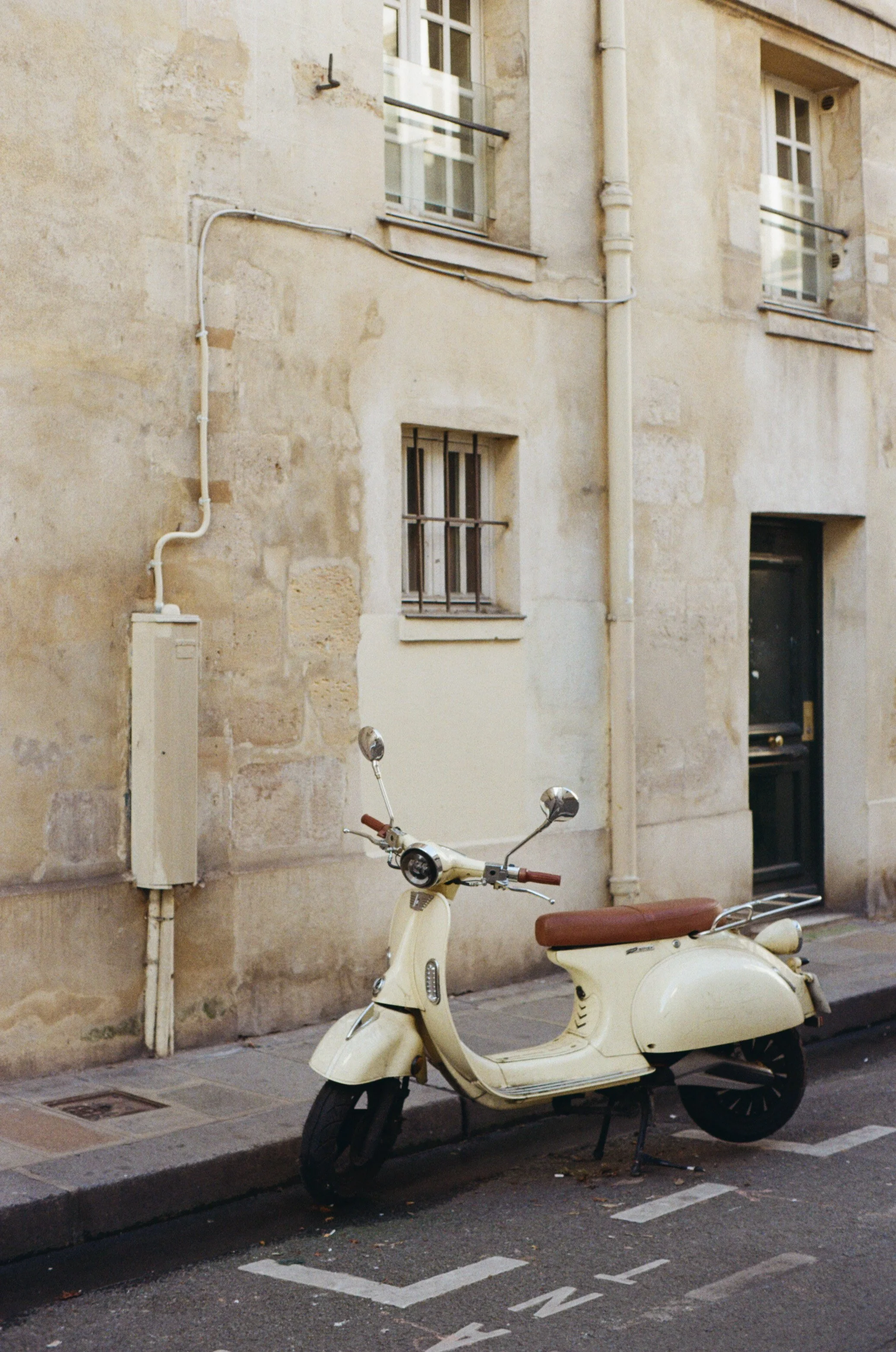 Scooter blanc stationné sur le trottoir devant un mur en pierre beige avec plusieurs fenêtres. Le mur montre des traces d'usure et des tuyaux. La voiture est dans un espace de stationnement nommé en blanc sur le sol.