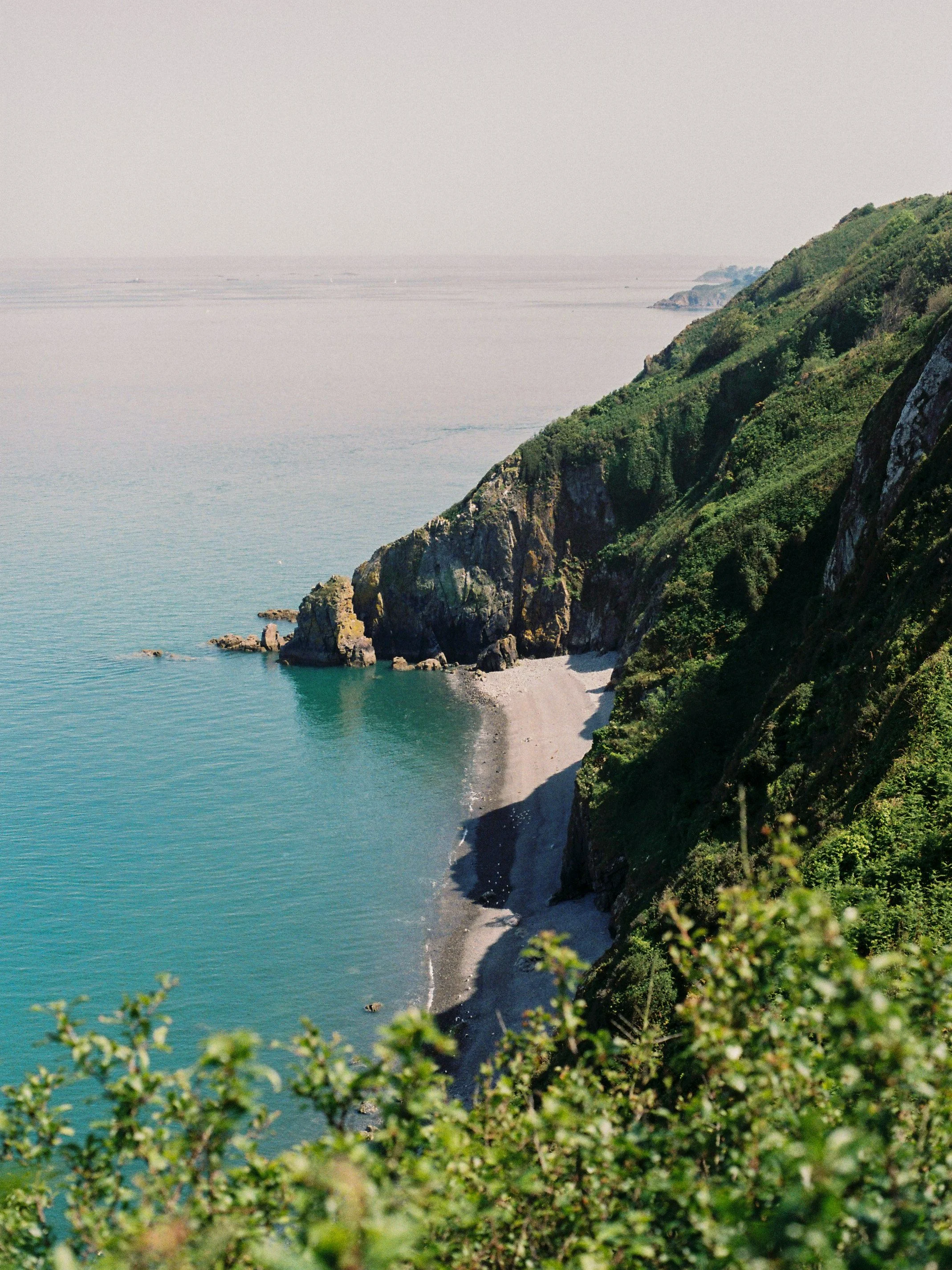 Crique rocheuse bordée d'eau turquoise et de falaises vertes, vue depuis un point élevé sur la côte, avec un ciel clair au loin.