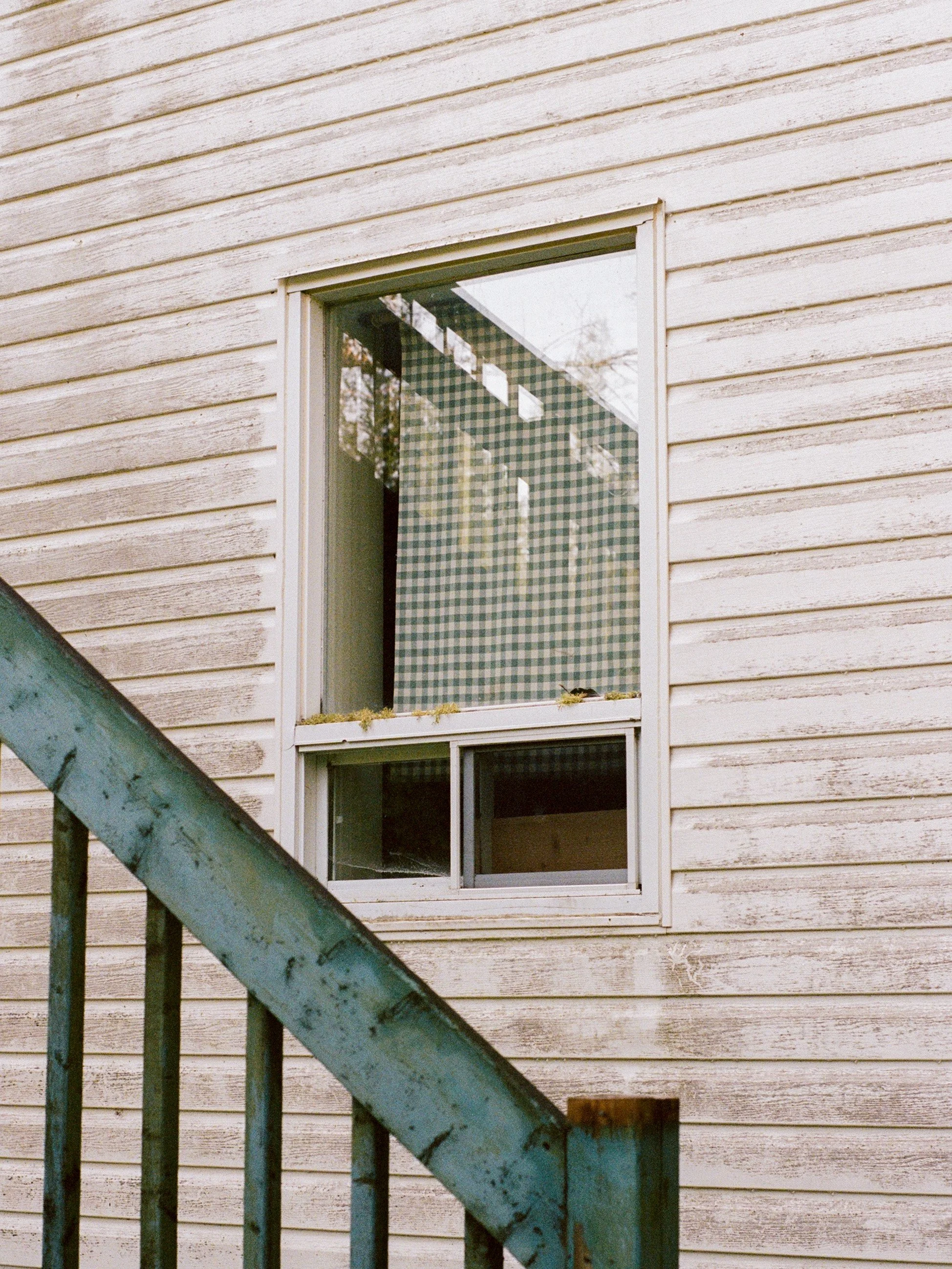 Fenêtre d'une maison en bois, vue de dessus, avec des cadres en PVC et un volet intérieur avec un motif à carreaux verts et blancs, et une échelle en métal rouillée à gauche.