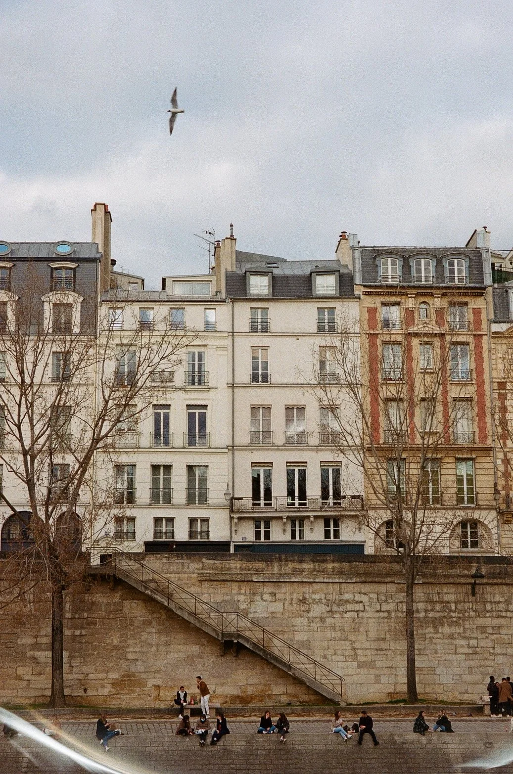 Un bâtiment résidentiel parisien vu depuis la Seine avec des arbres nus, des personnes assises sur les marches et un oiseau volant dans le ciel nuageux.