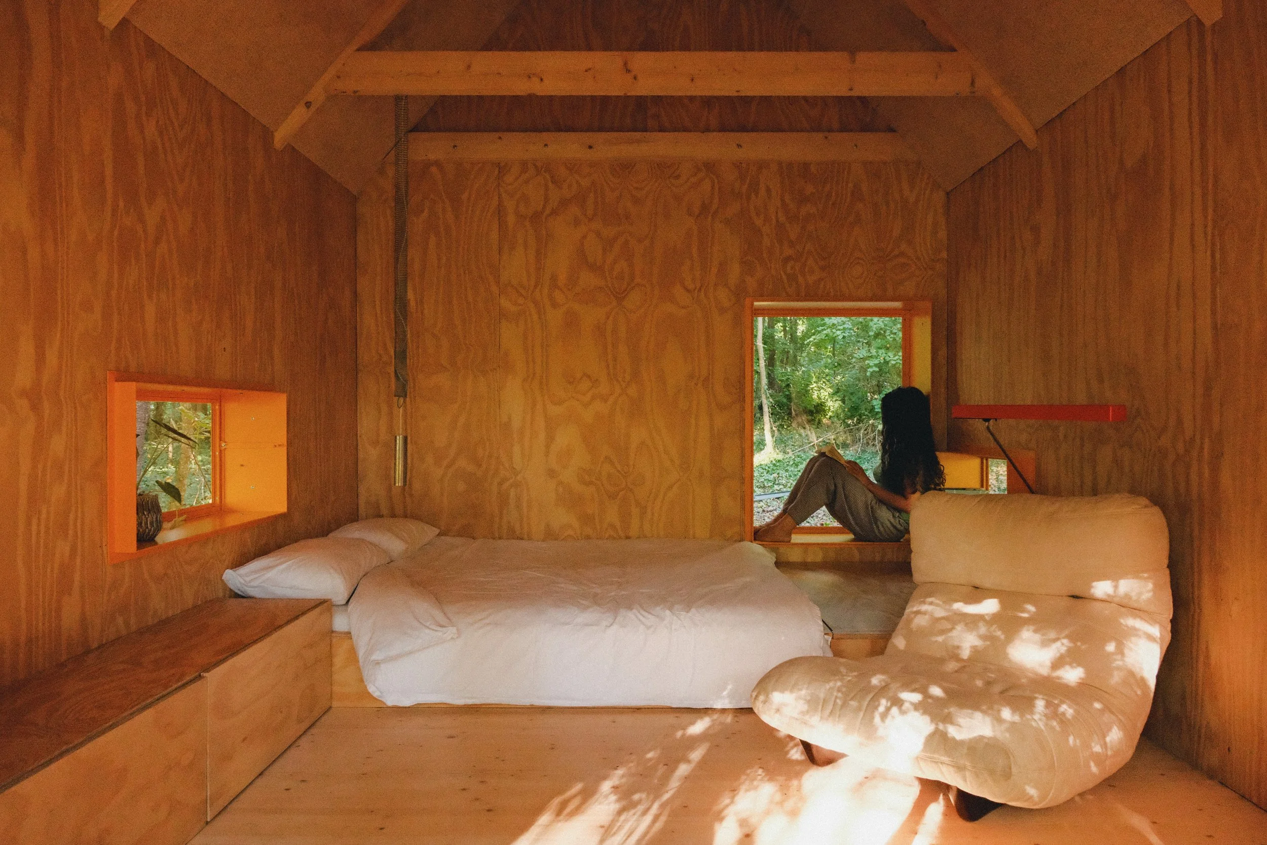 Une chambre en bois avec deux fenêtres laissant entrer la lumière naturelle, une personne assise sur un rebord de fenêtre regardant à l'extérieur, et un lit avec des draps blancs.