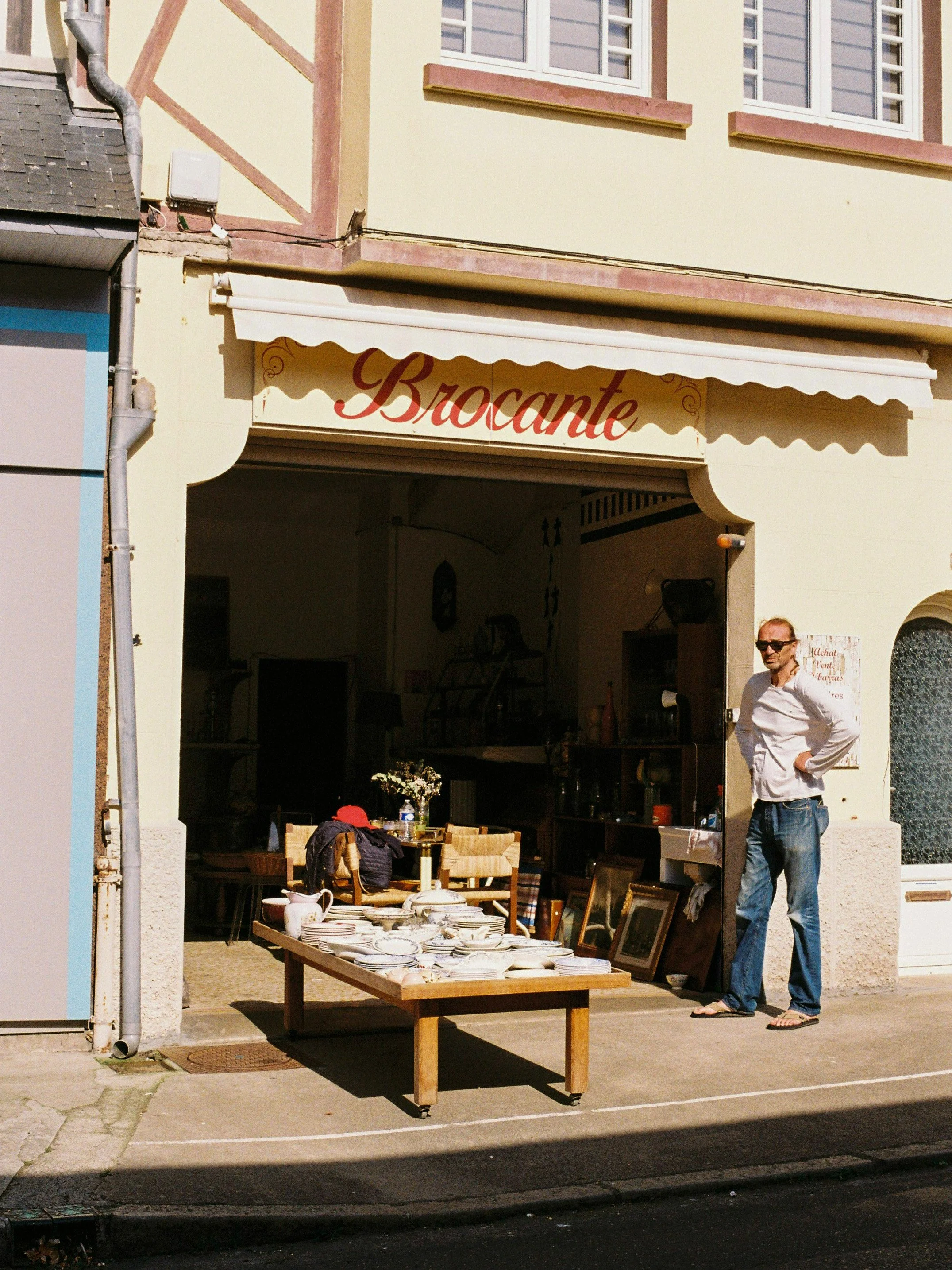 Une personne se tient devant une boutique ou un marché avec des objets en vente sur une table. La devanture de la boutique porte le nom 'Bracante'.