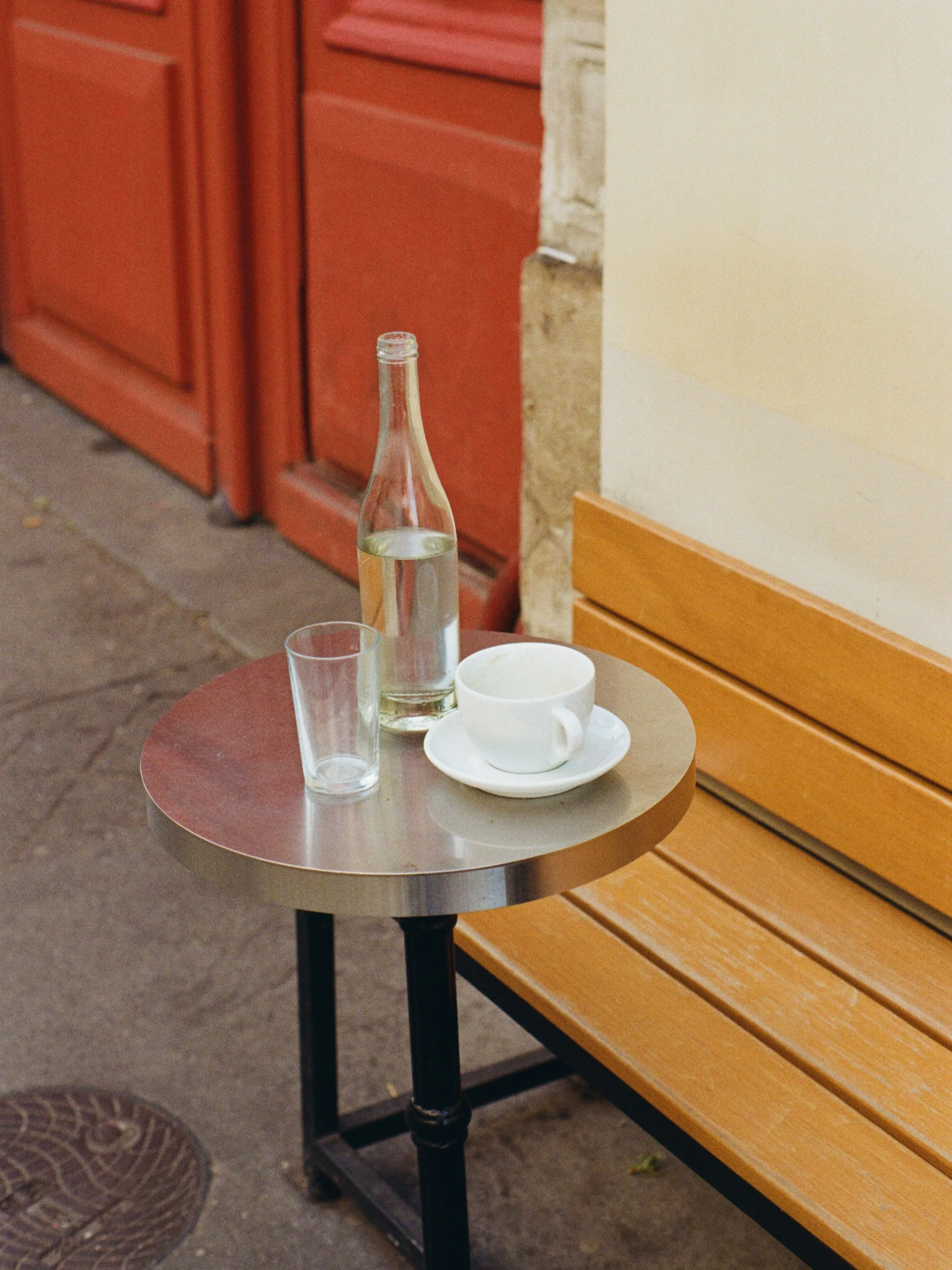 Une petite table ronde en métal avec une bouteille d'eau, un verre vide, un café blanc et une assiette blanche, en face d'un banc en bois dans un intérieur ou un café.