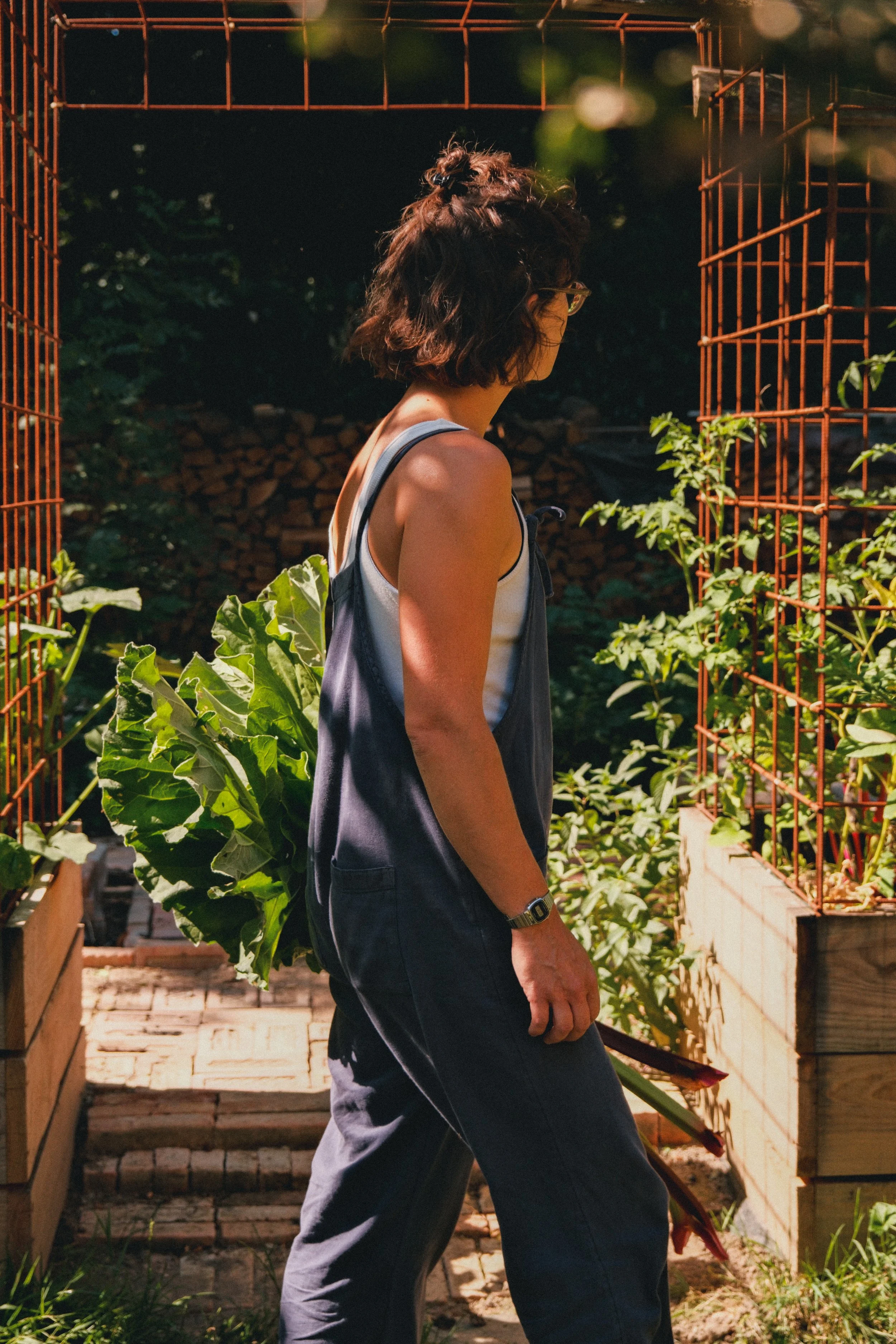 Une personne vue de profil dans un jardin, tenant un bouquet de fleurs, entourée de plantes vertes et d'une structure en métal rouge.
