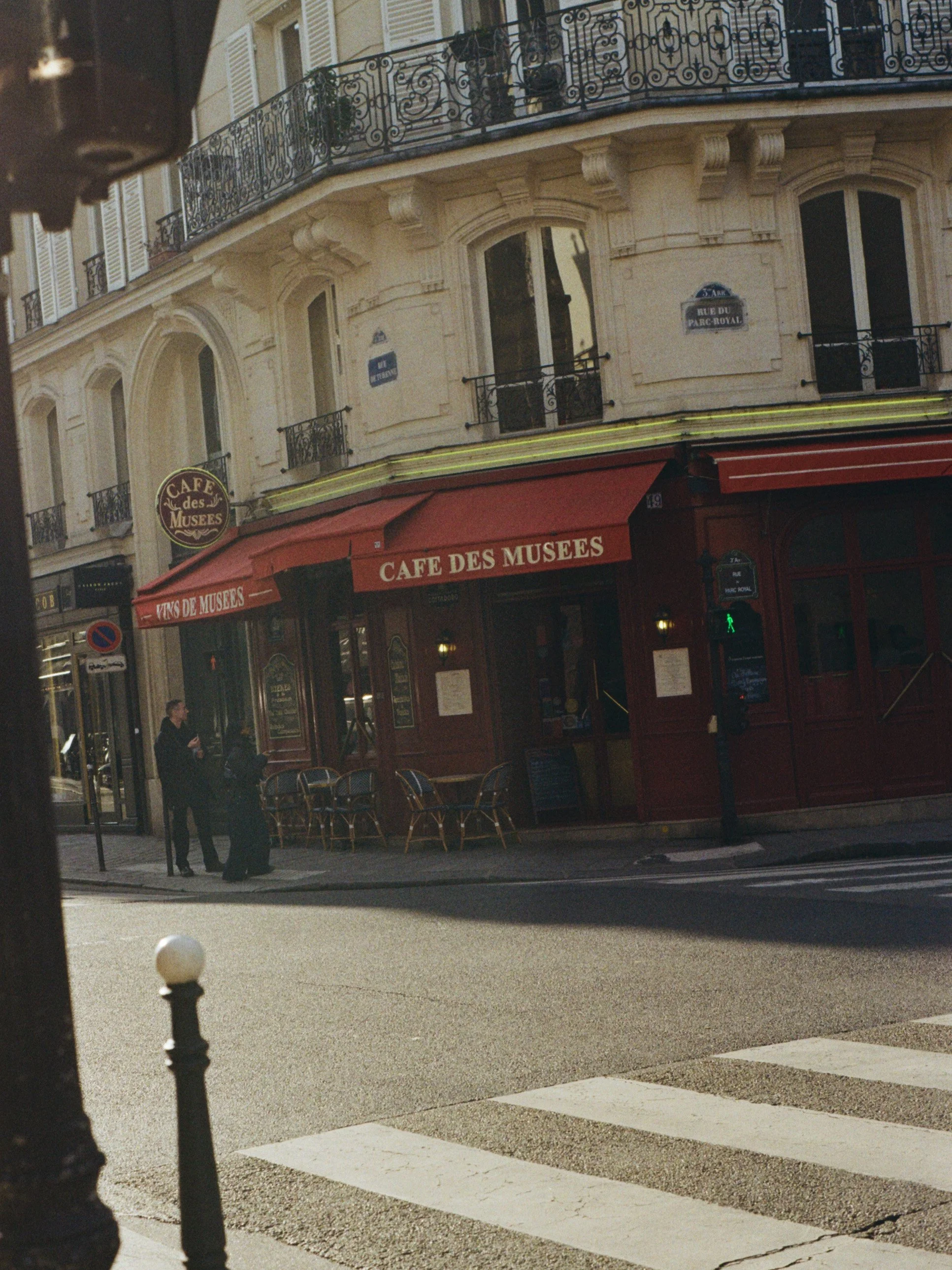 Une rue parisienne avec un bâtiment en architecture classique, une enseigne 'Café des Musées' sous une veranda rouge, et un passage piéton. Deux personnes sont visibles à l'arrêt au coin de la rue.