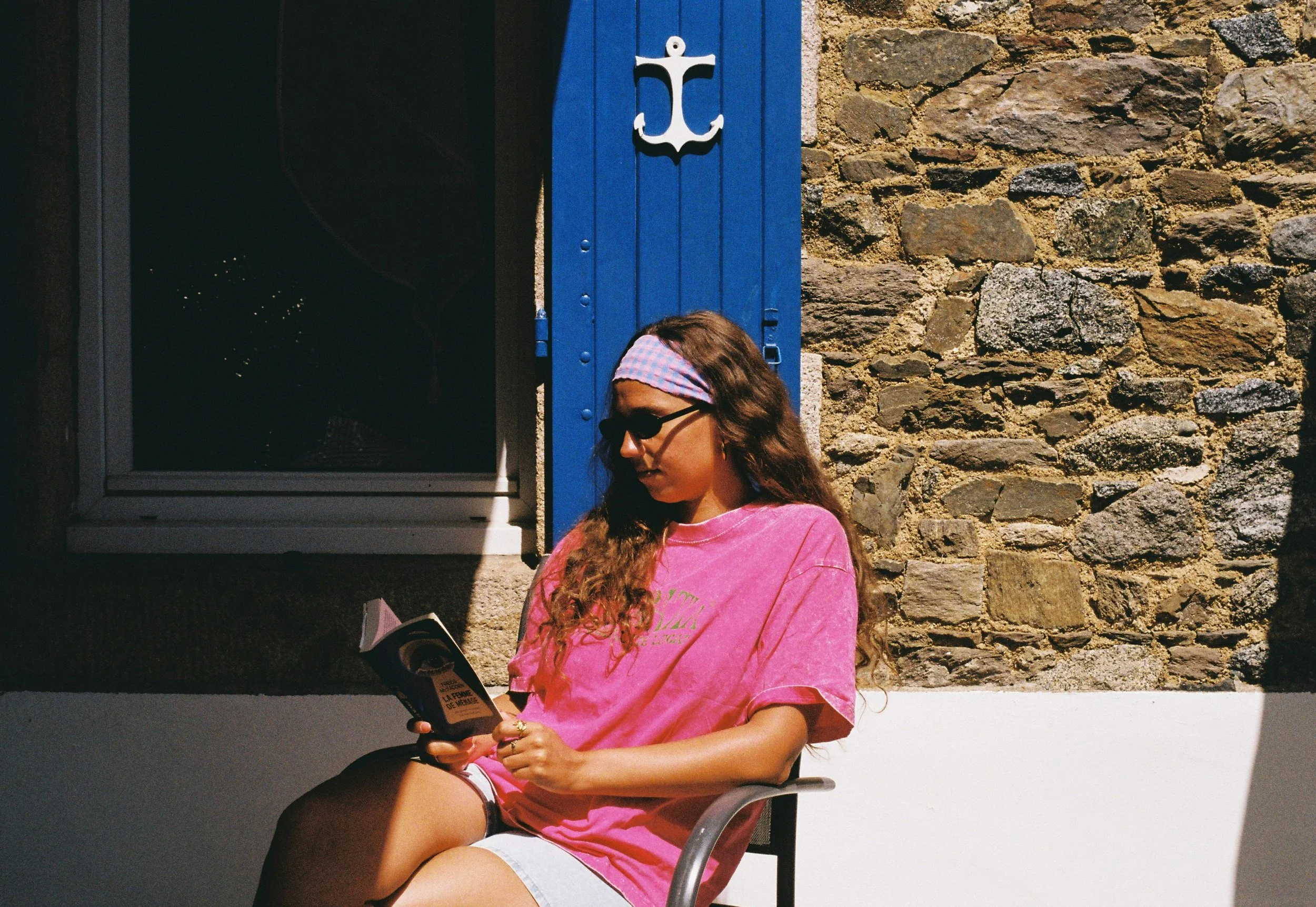 Une jeune femme portant des lunettes noires, un bandeau rose et une chemise rose, assise en plein air et lisant un livre, avec un mur en pierre derrière elle.