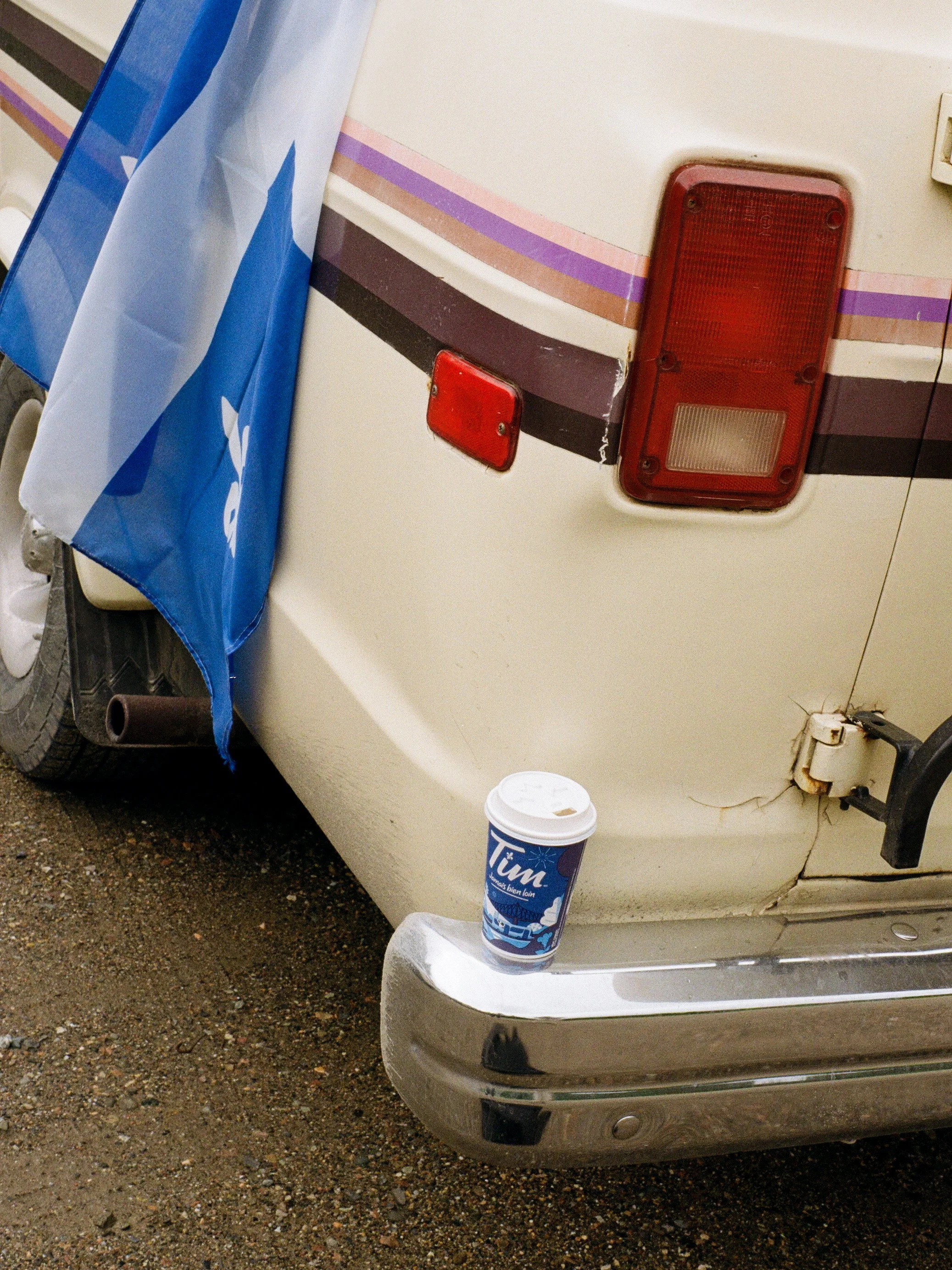Une vieille voiture beige avec des rayures roses et violettes, et deux feux arrières rouges, stationnée sur un sol en terre. Un gobelet en plastique blanc avec une inscription bleue est posé sur la porte de la voiture.